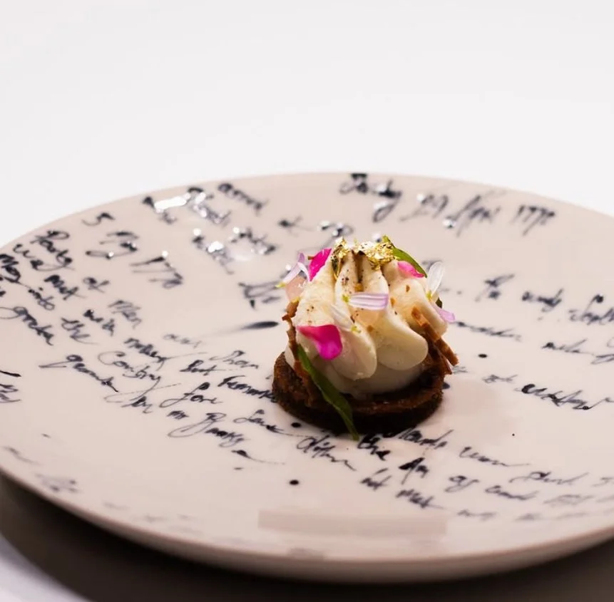A decorative plate with handwritten script design holding a small gourmet appetizer topped with flowers and edible decorations.