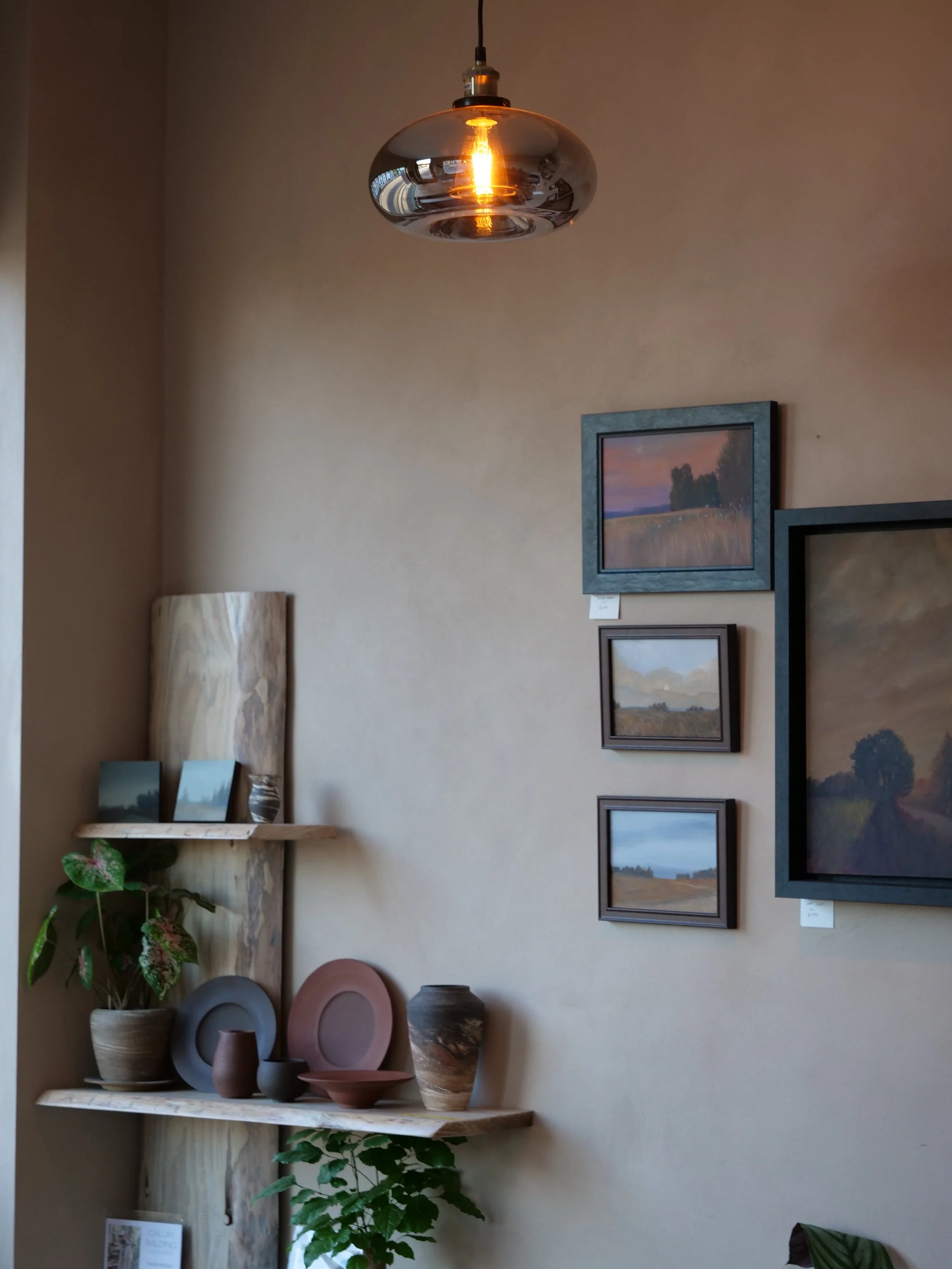 Interior art gallery with framed landscape paintings on the wall, a wood shelf with decorative pottery and pictures, a plant, and a modern pendant light hanging from the ceiling.