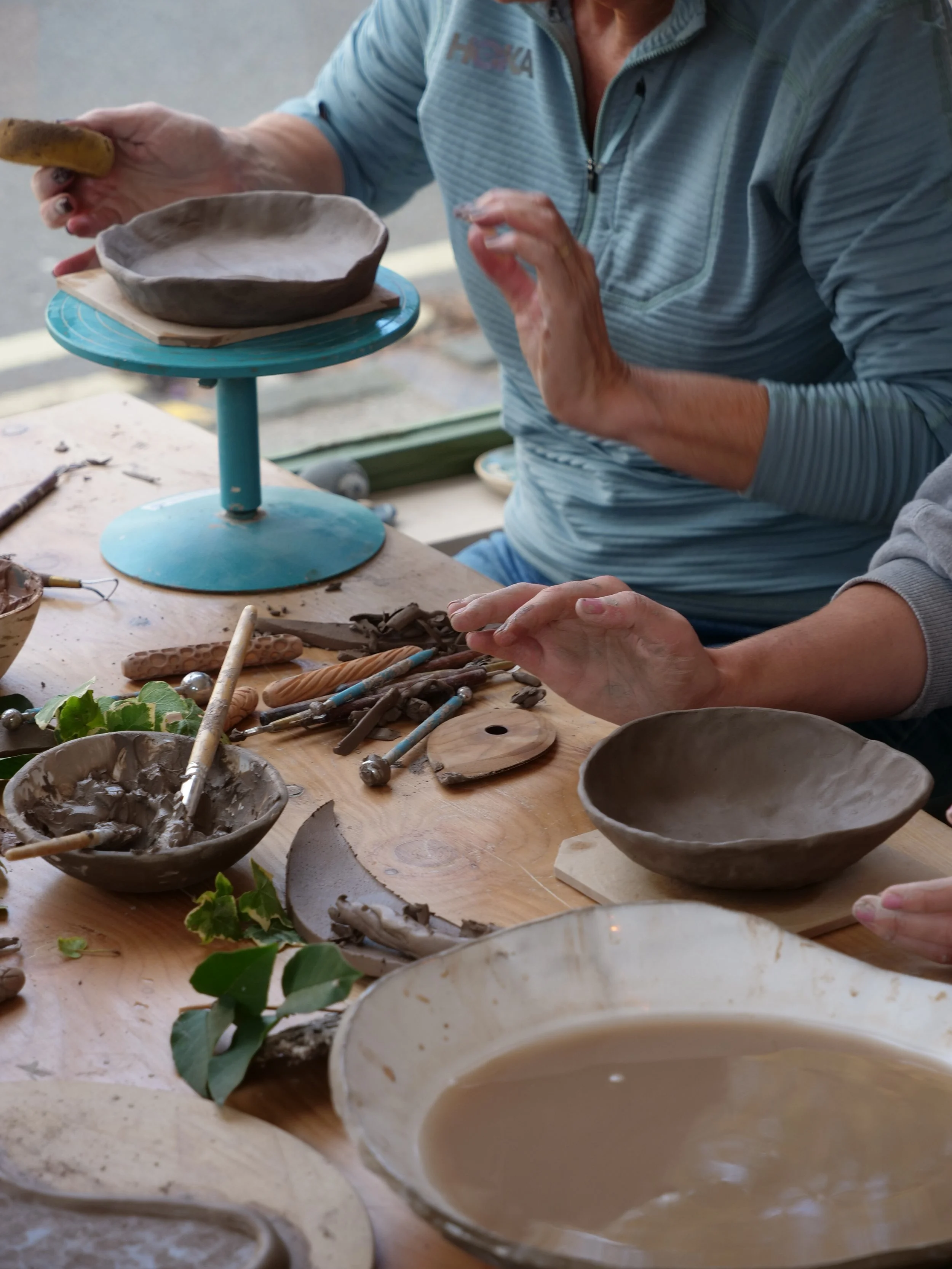 Two people working on pottery at a craft workshop, with bowls, clay tools, and clay pieces on the table.