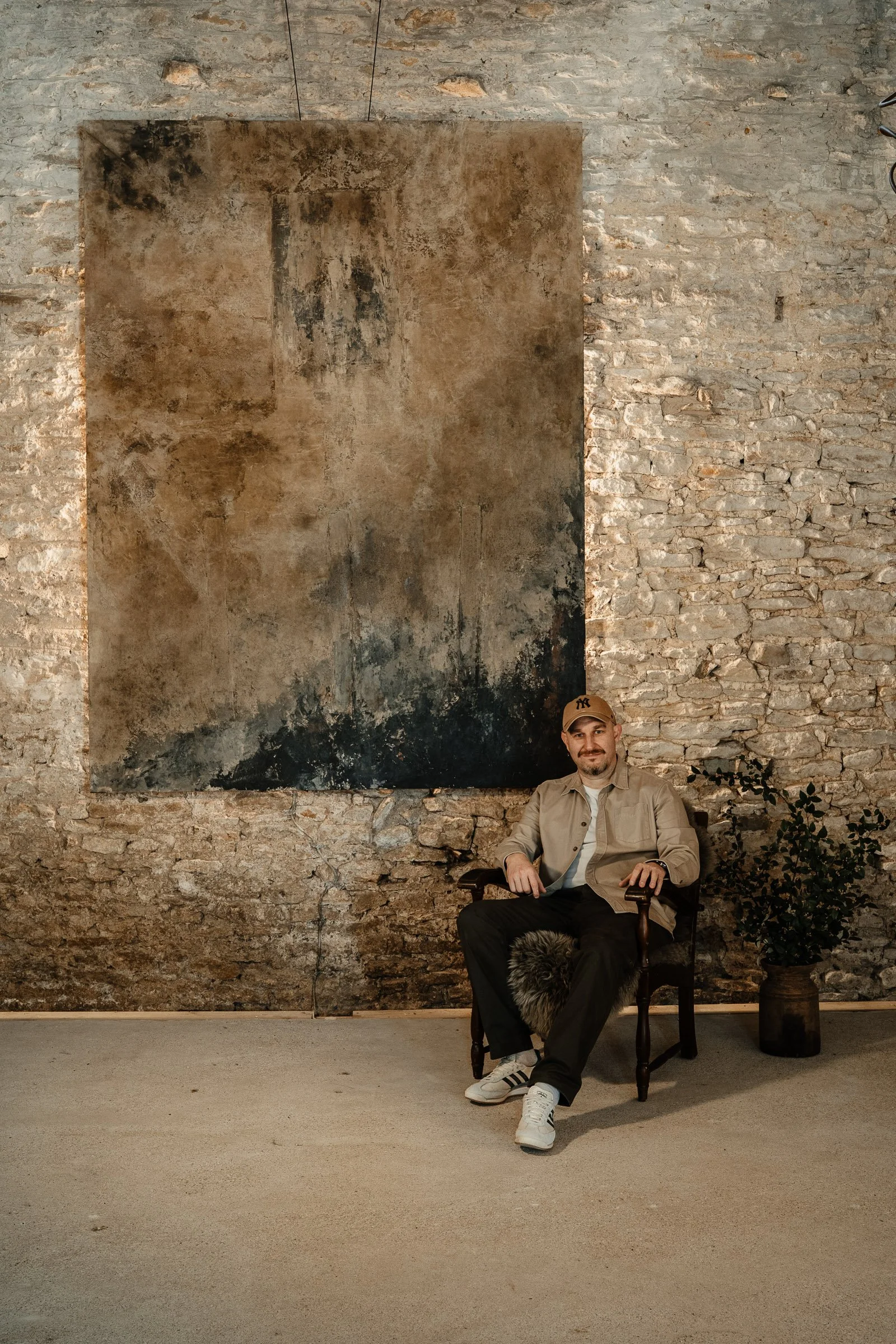 A man sitting on a wooden chair with a fur pillow, in front of a brick wall with an abstract painting, and a potted plant to his right.