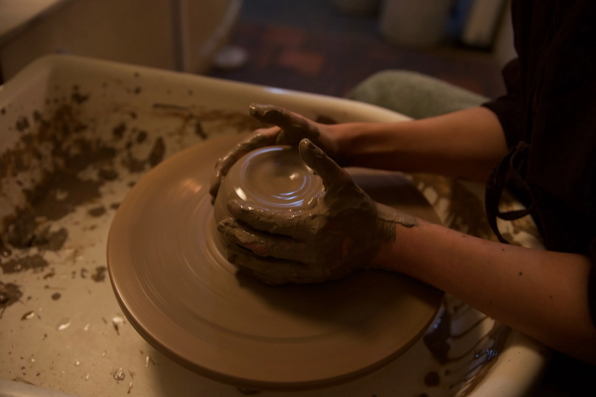 Person shaping clay on a pottery wheel in a dimly lit pottery studio.