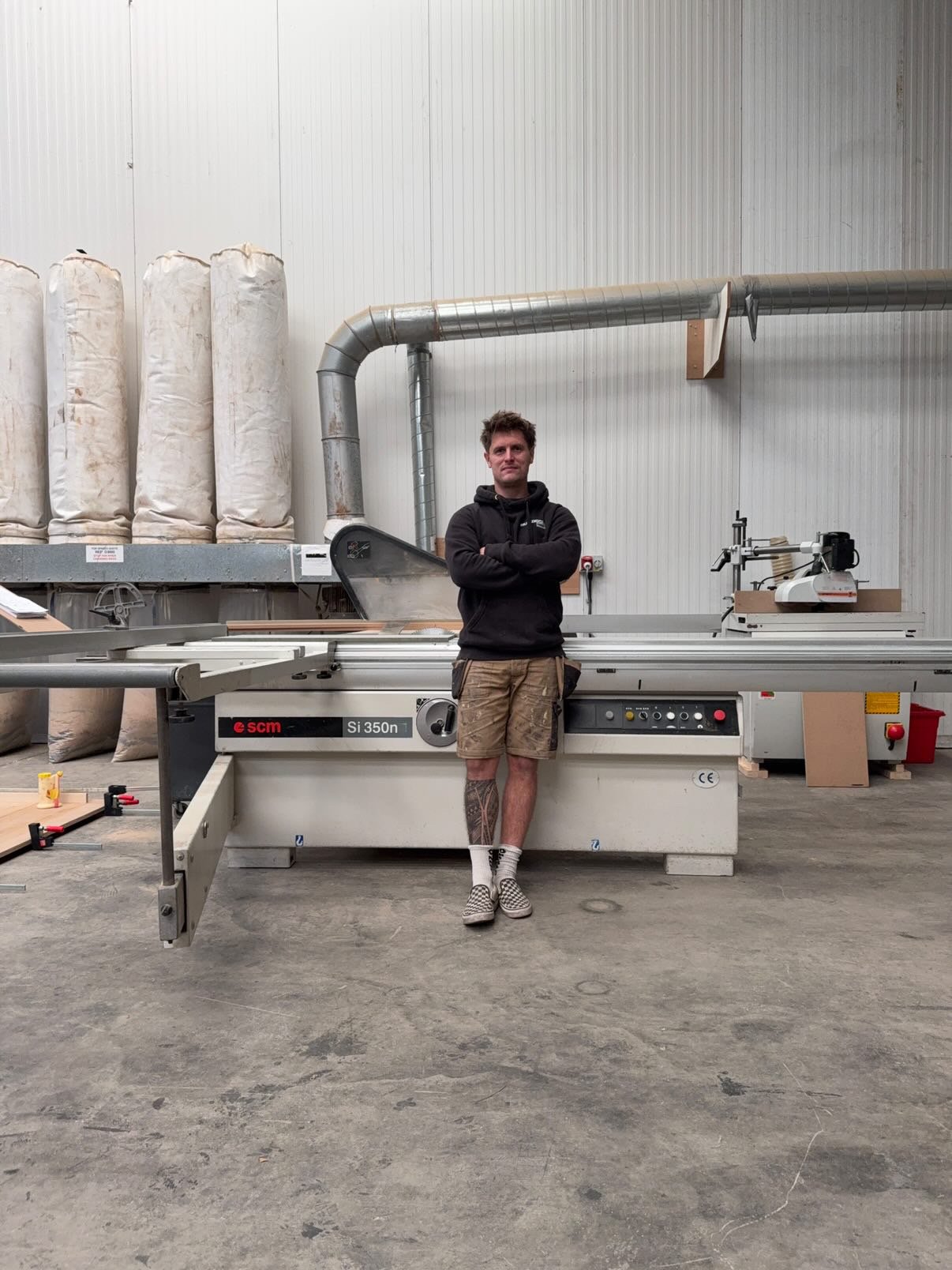 A man standing with his arms crossed in front of a large industrial woodworking machine in a workshop. The man is wearing a black hoodie, shorts, and checkerboard slip-on shoes, with tattoos visible on his left leg.