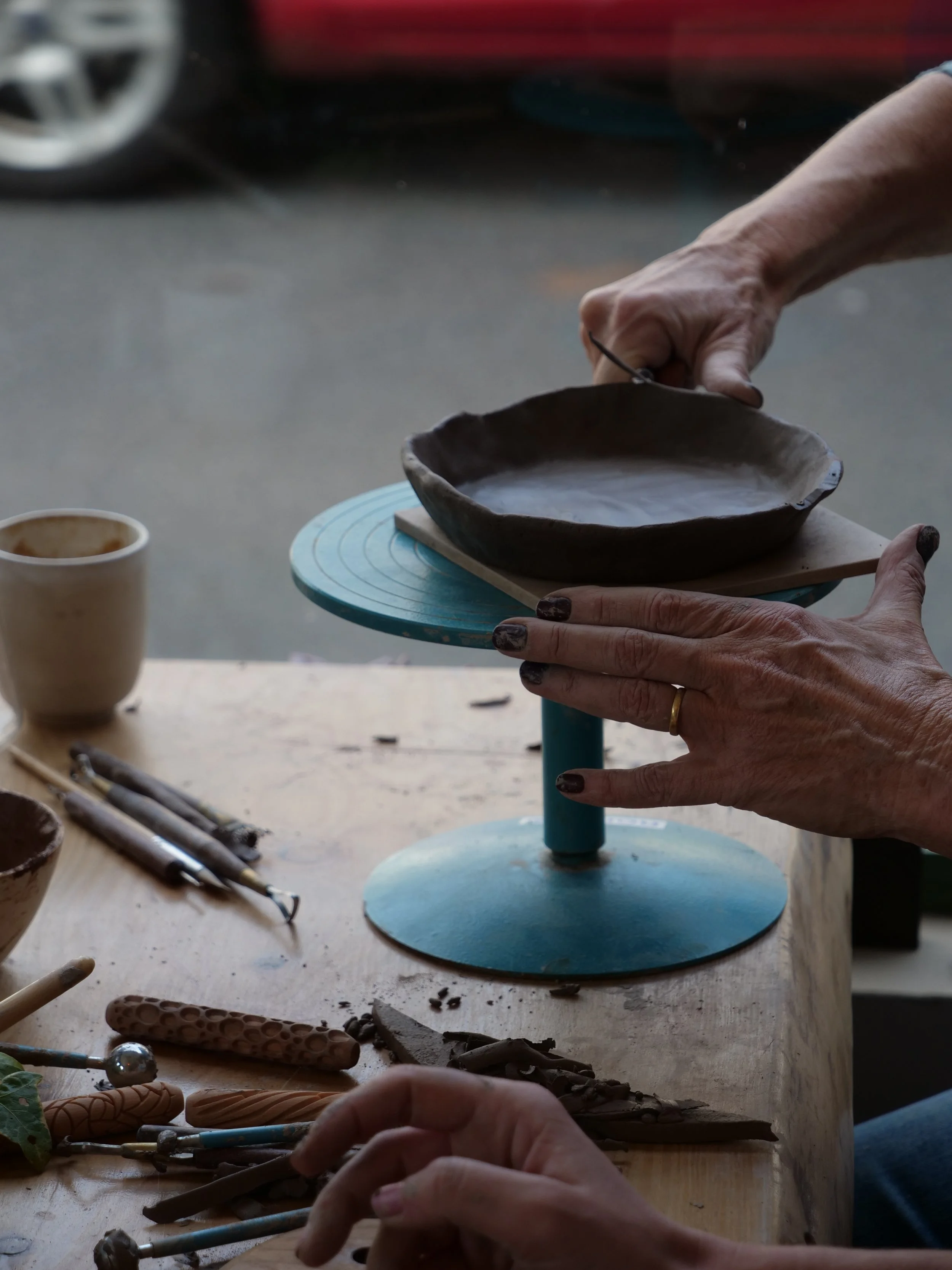 Person shaping a clay dish on a small blue turntable with pottery tools and a cup nearby.