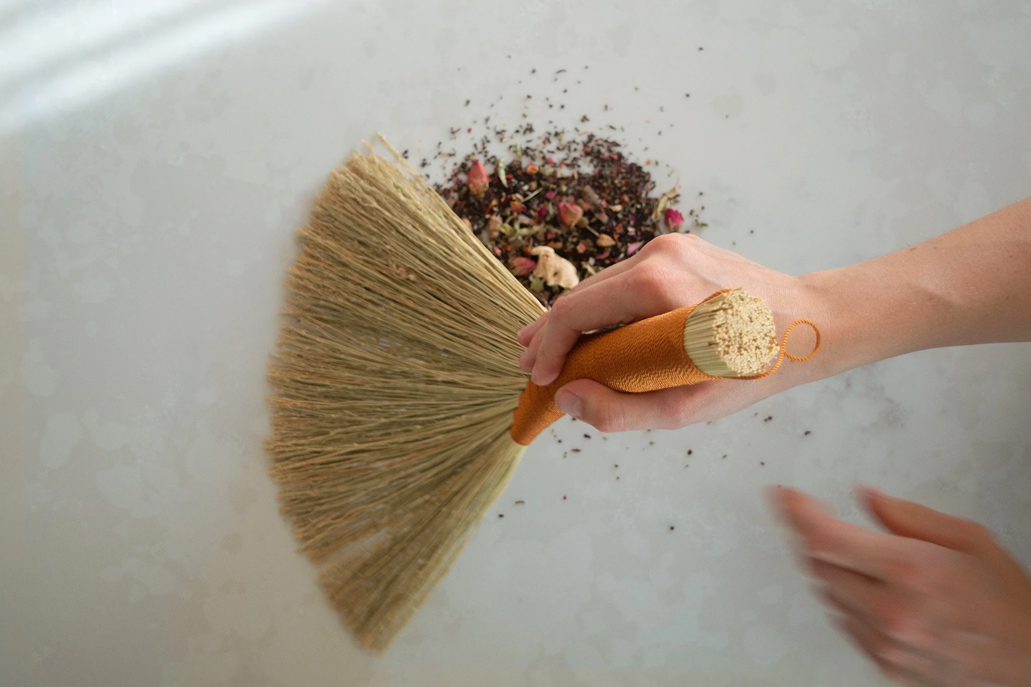 A hand holding a small broom sweeping up tea leaves and dried rose petals on a white surface.