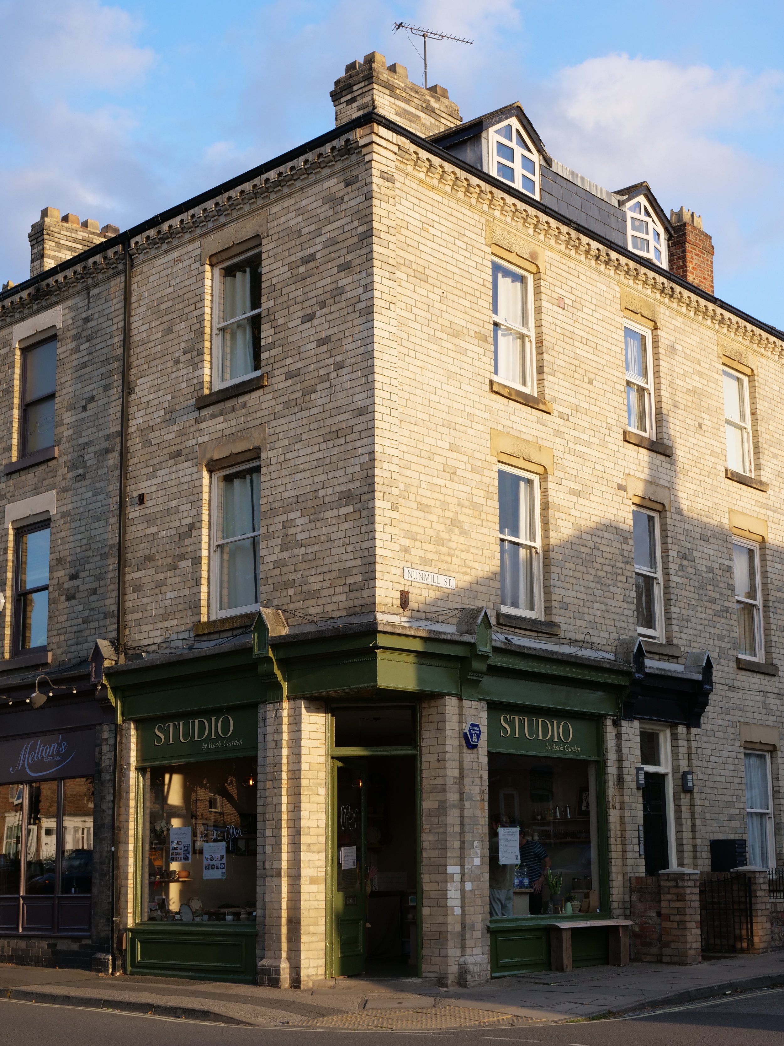 A corner view of a three-story brick building with a green storefront labeled "STUDIO by Rock Garden," located at the intersection of unmarked streets. The building has multiple windows, some with curtains, and a small sign with street name "Unummill