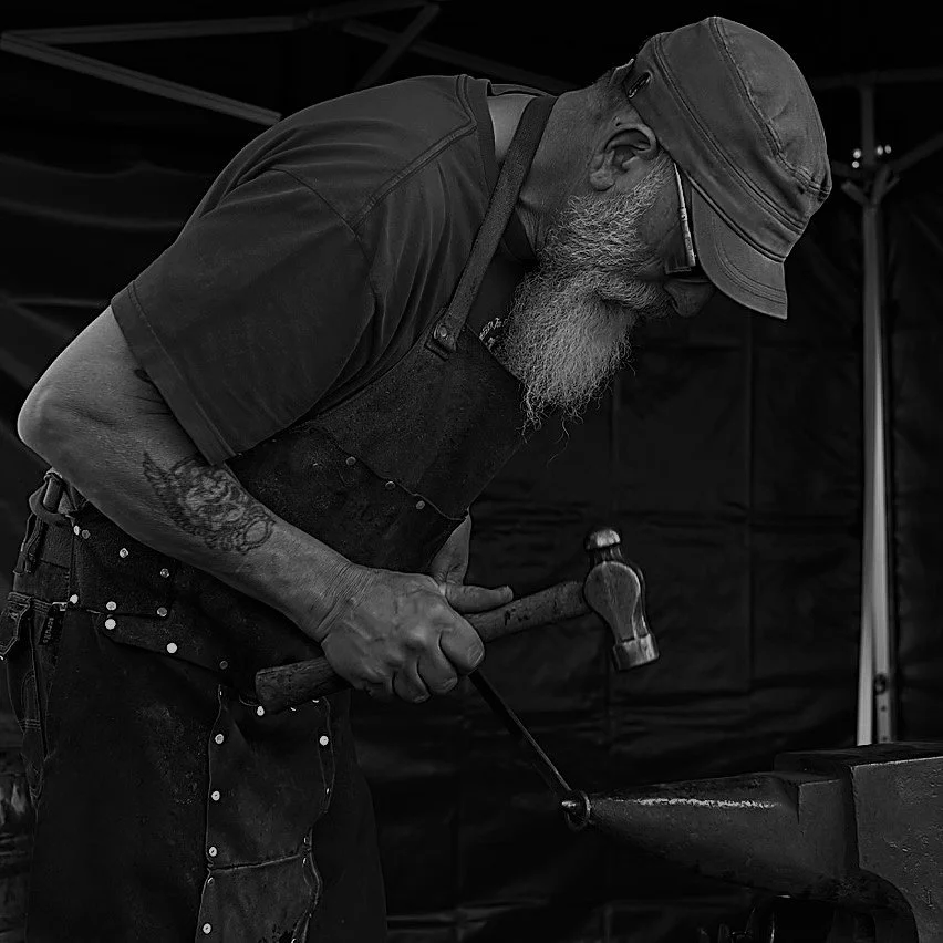 An older man with a beard wearing glasses, a hat, and an apron, shaping metal on an anvil with a hammer in a black and white photo.