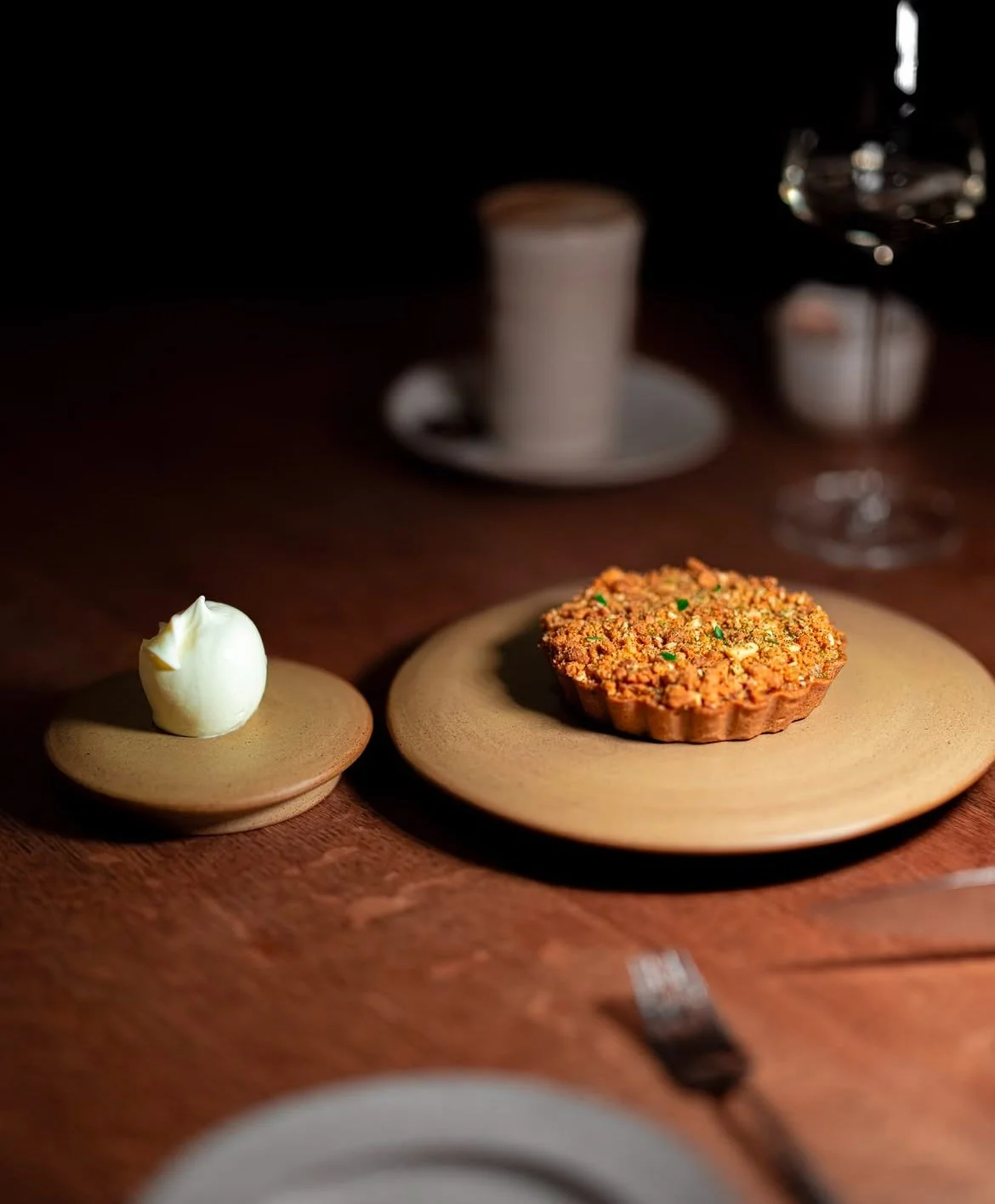 A dessert on a wooden plate, a small dollop of whipped cream on a smaller plate, a glass of red wine, a candle, and a cup in the background on a dark wooden table.
