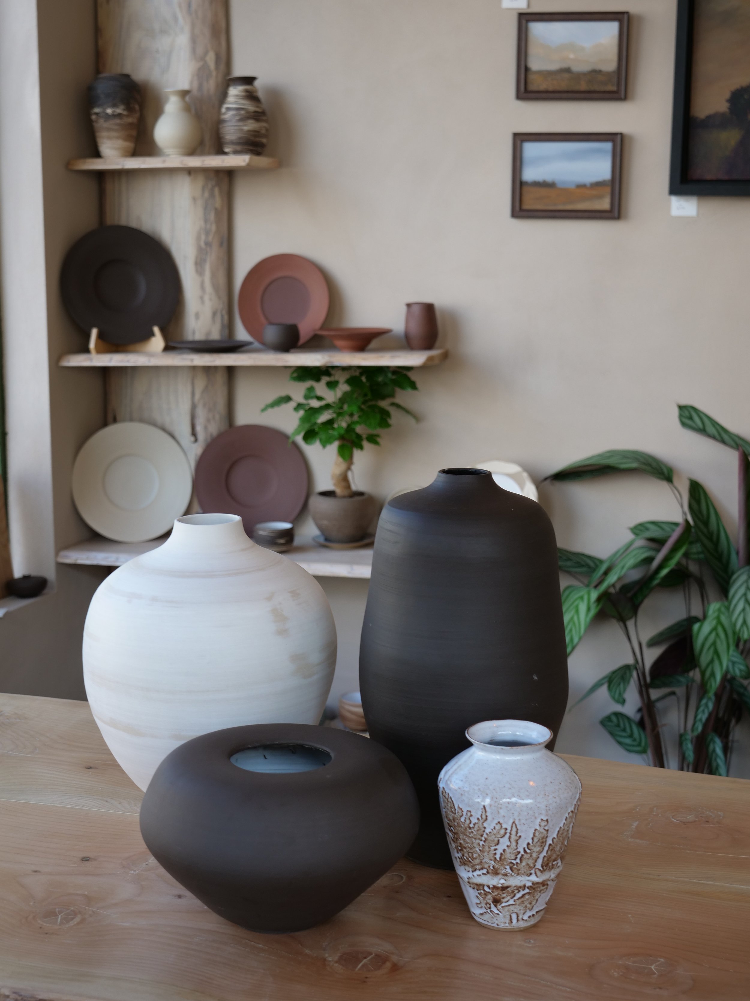 Five vases of different sizes, colors, and shapes on a wooden table, with a background of shelves holding pottery and framed landscape paintings.