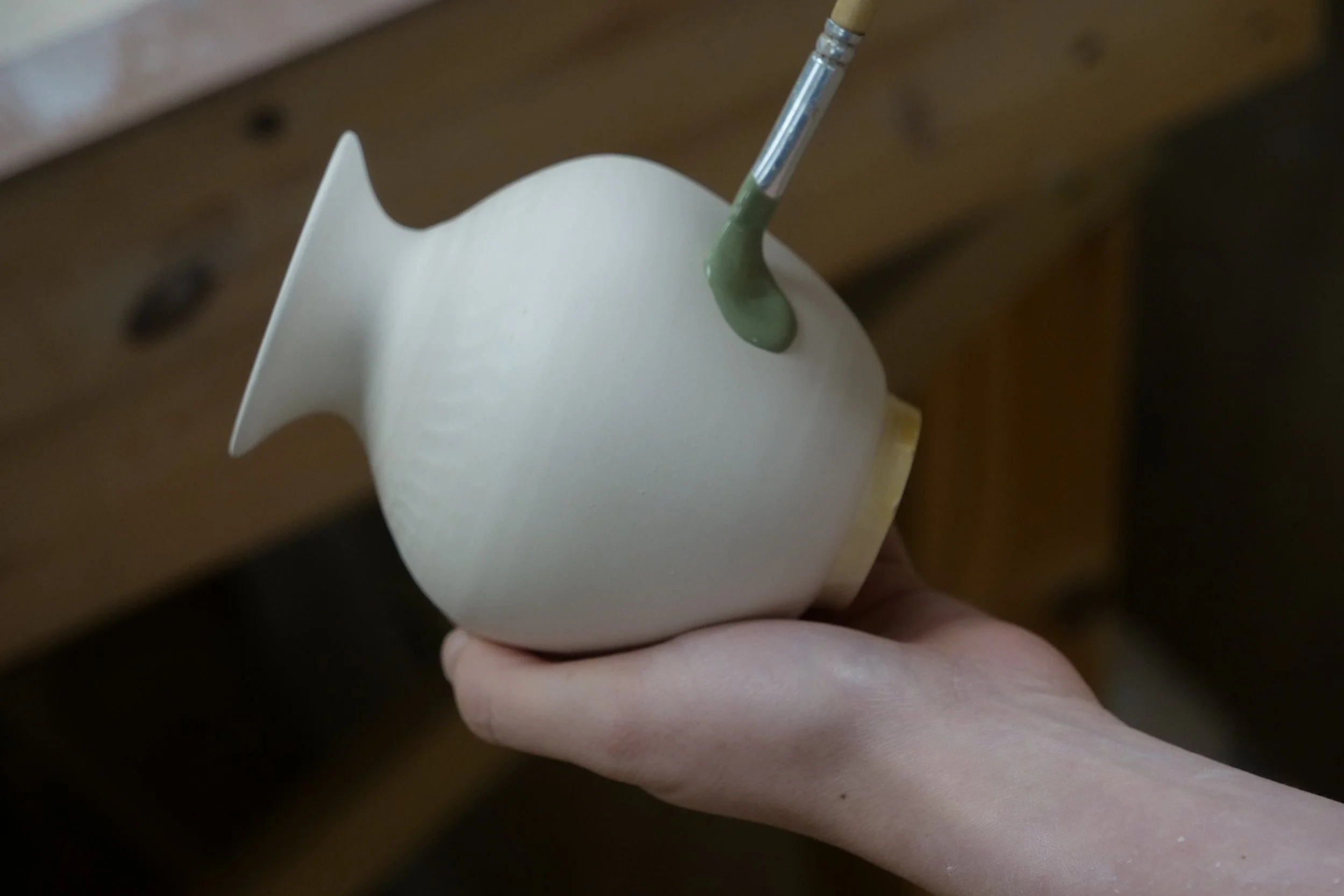 A person holding a ceramic fish-shaped planter with a paintbrush applying green glaze to it.