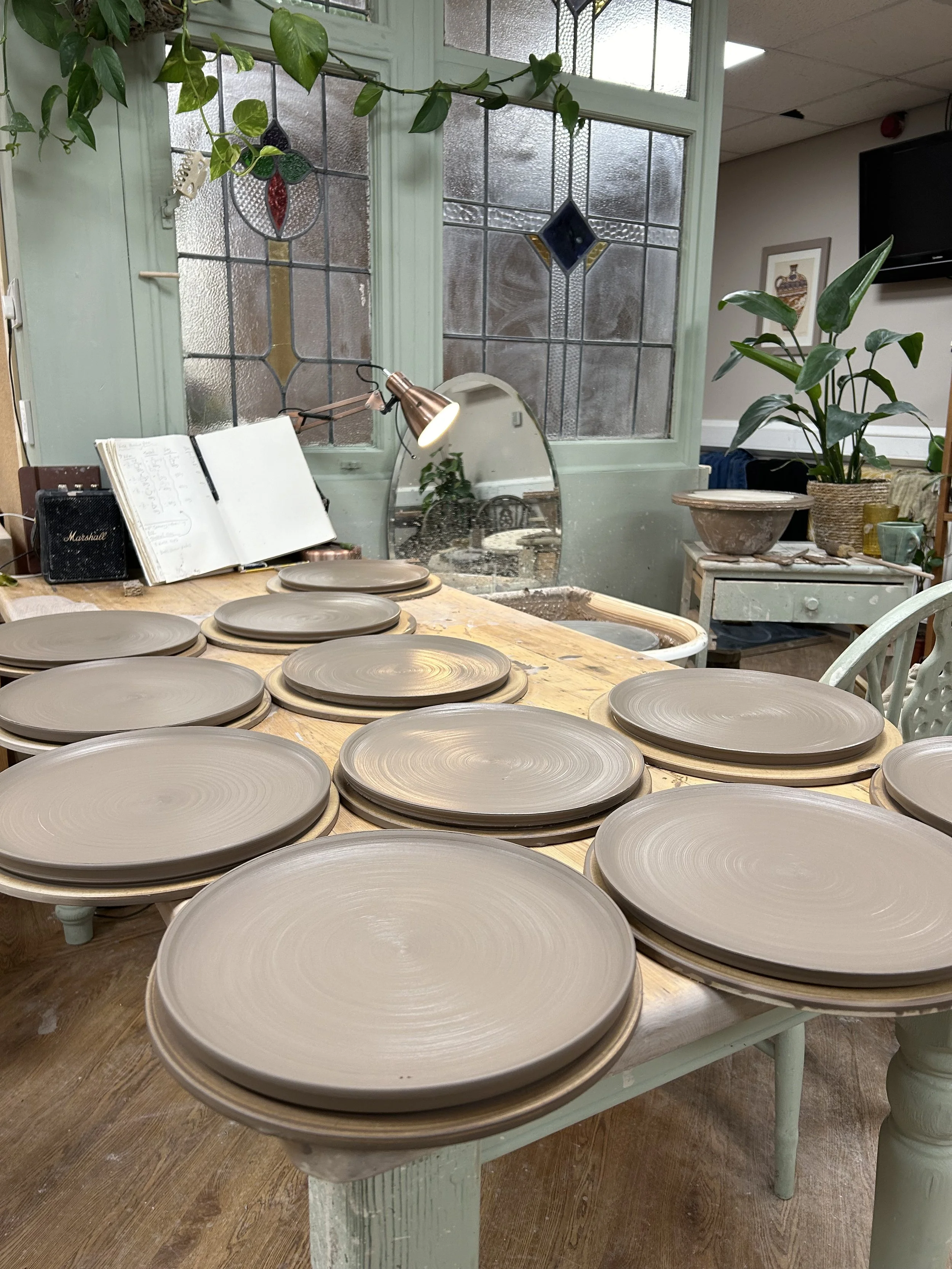 A dining table with multiple plain, unglazed ceramic plates stacked in clockwise formation, with a mirror and decorative stained glass window in the background, and potted plants on side tables.