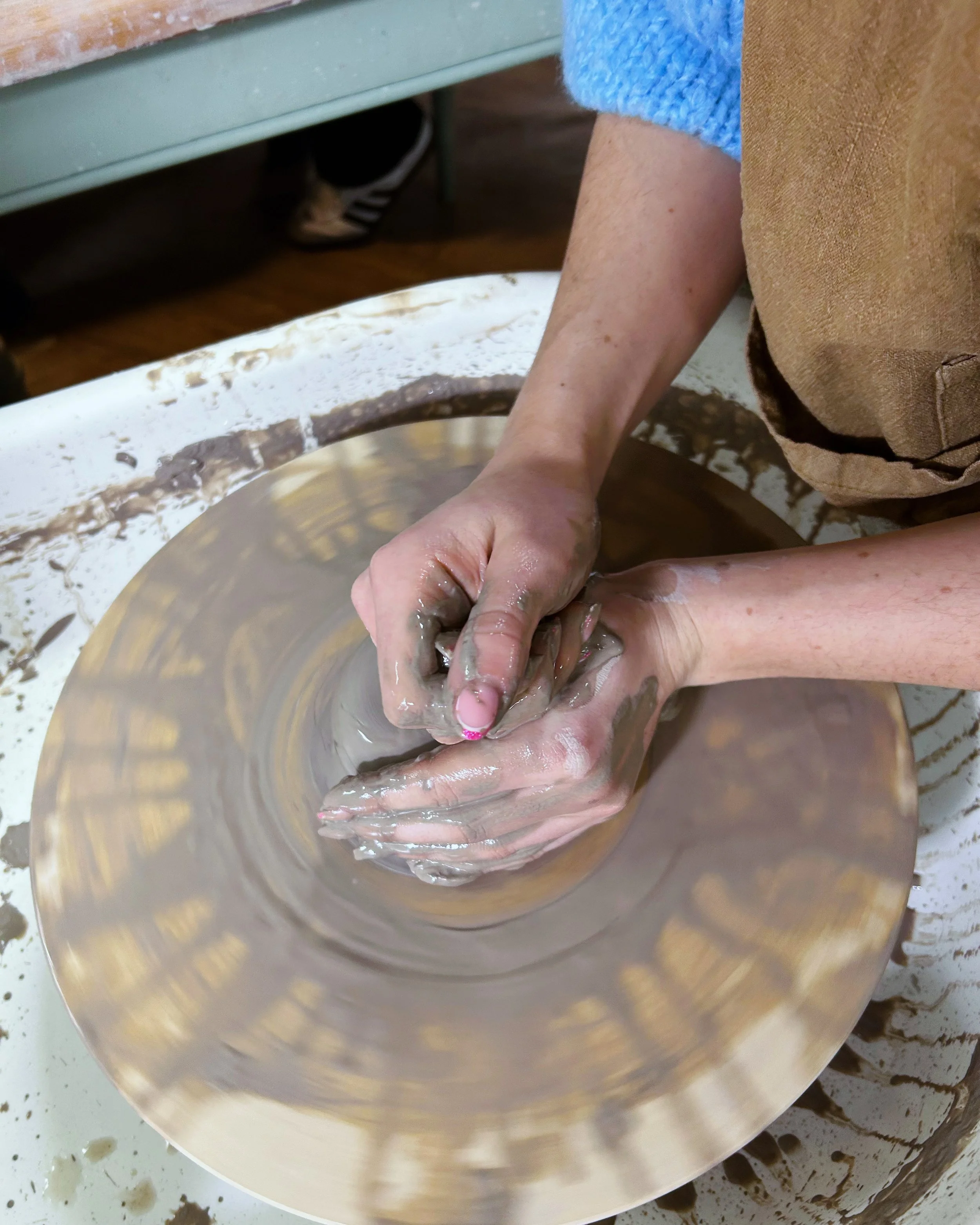 Two hands shaping clay on a pottery wheel inside a room.