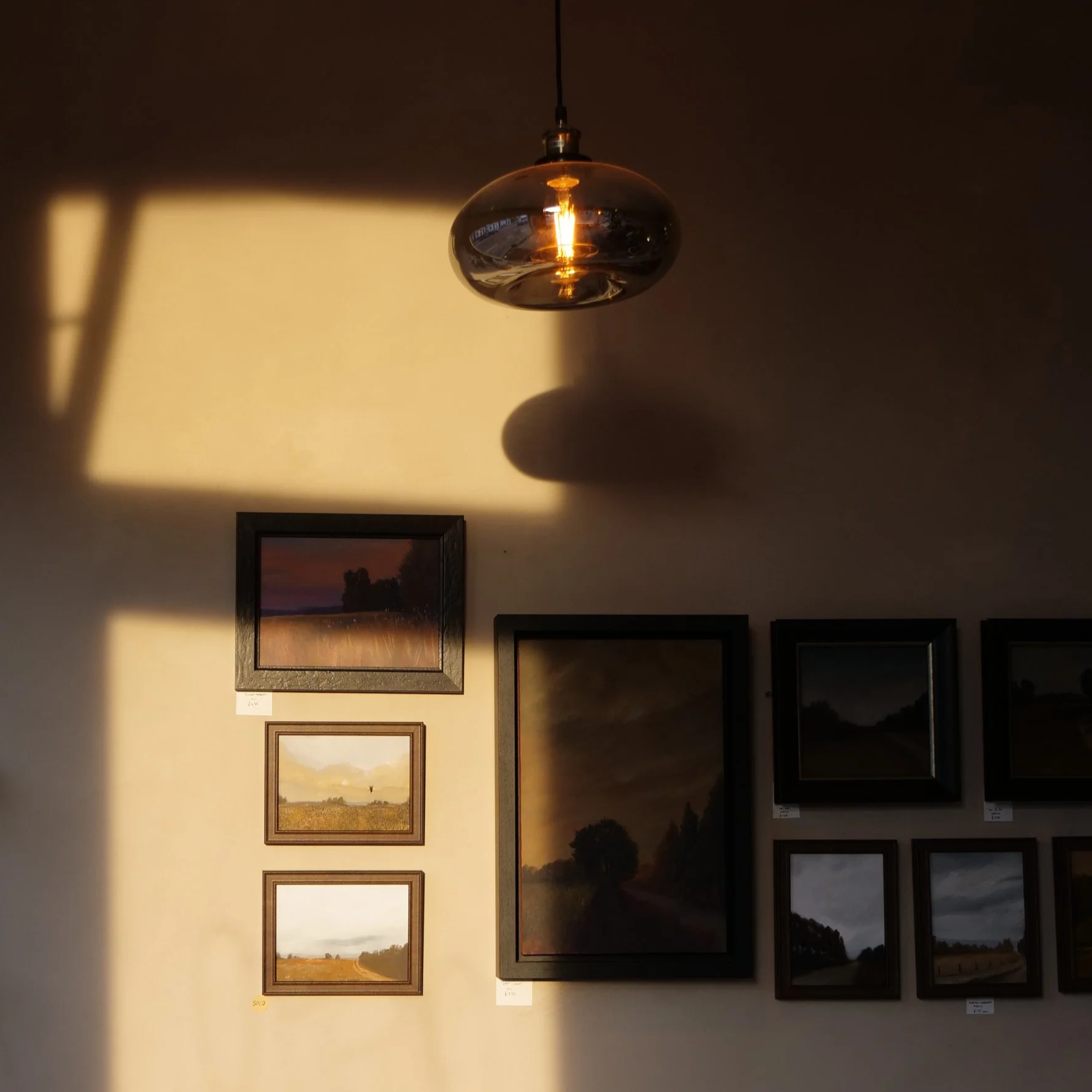 Art gallery wall with framed landscape paintings and a hanging pendant light casting shadows on the wall.