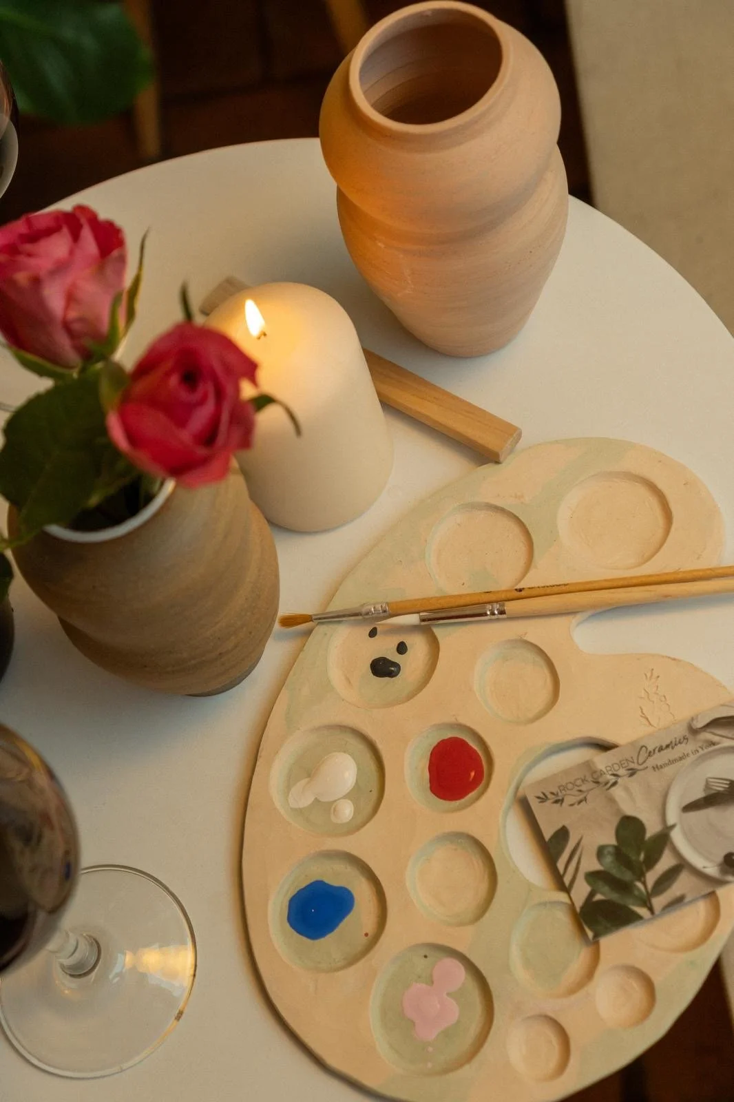 A table with pink roses in a small vase, a lit white candle, wooden vases, and a wooden paint palette with various colors of paint and a paintbrush.