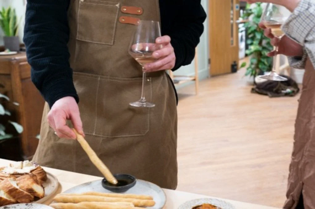 A person wearing a brown apron dips breadsticks into a black dipping bowl with a glass of rosé wine in their other hand, standing near a table with baked goods in a cozy indoor setting.