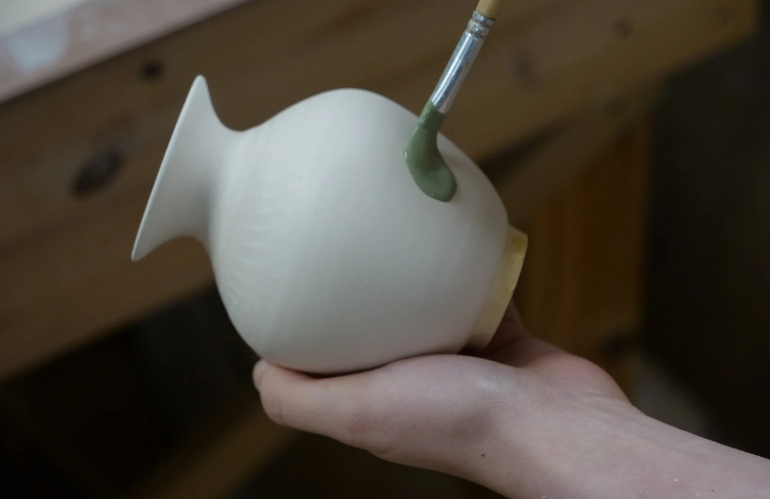 Person is holding a small, white ceramic fish-shaped vessel with a hole at the top, from which green glaze is being poured with a brush.