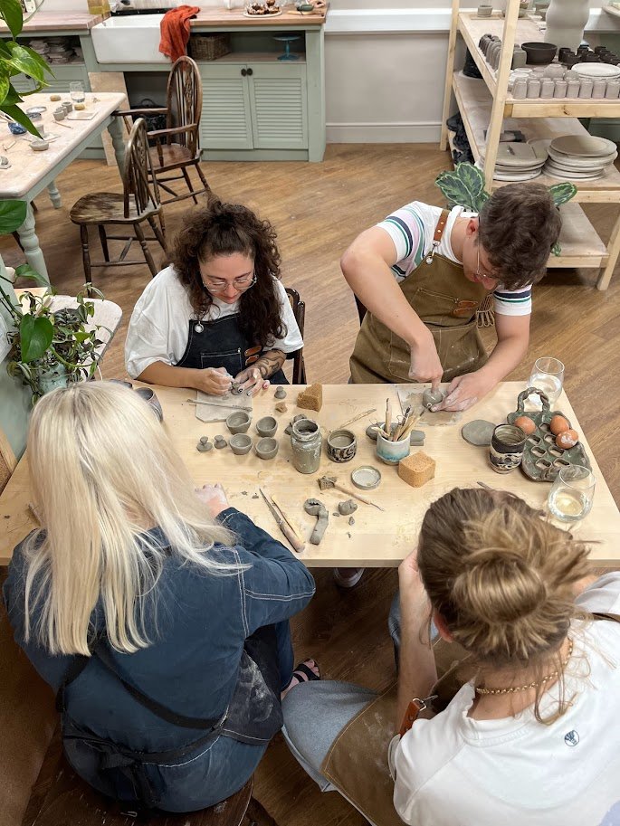 People participating in a pottery class, working on clay projects at a wooden table in a cozy studio.