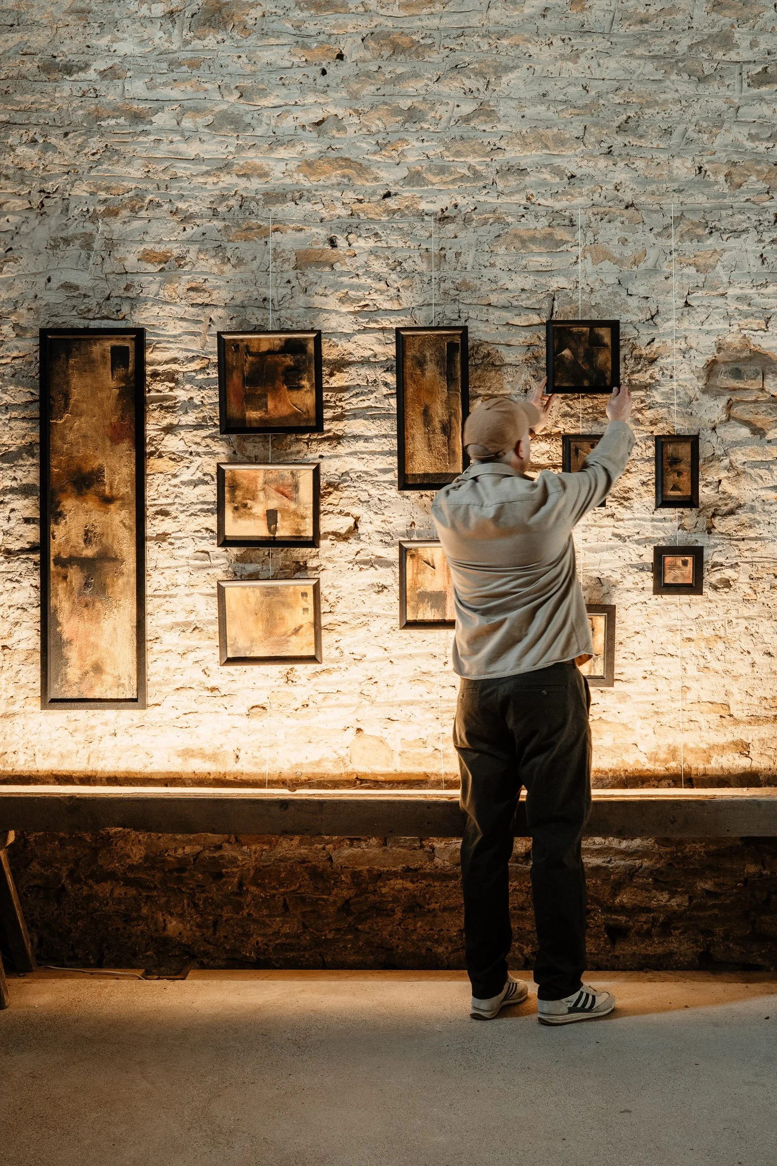 A person standing in an art gallery, adjusting or hanging framed abstract artwork on a brick wall.
