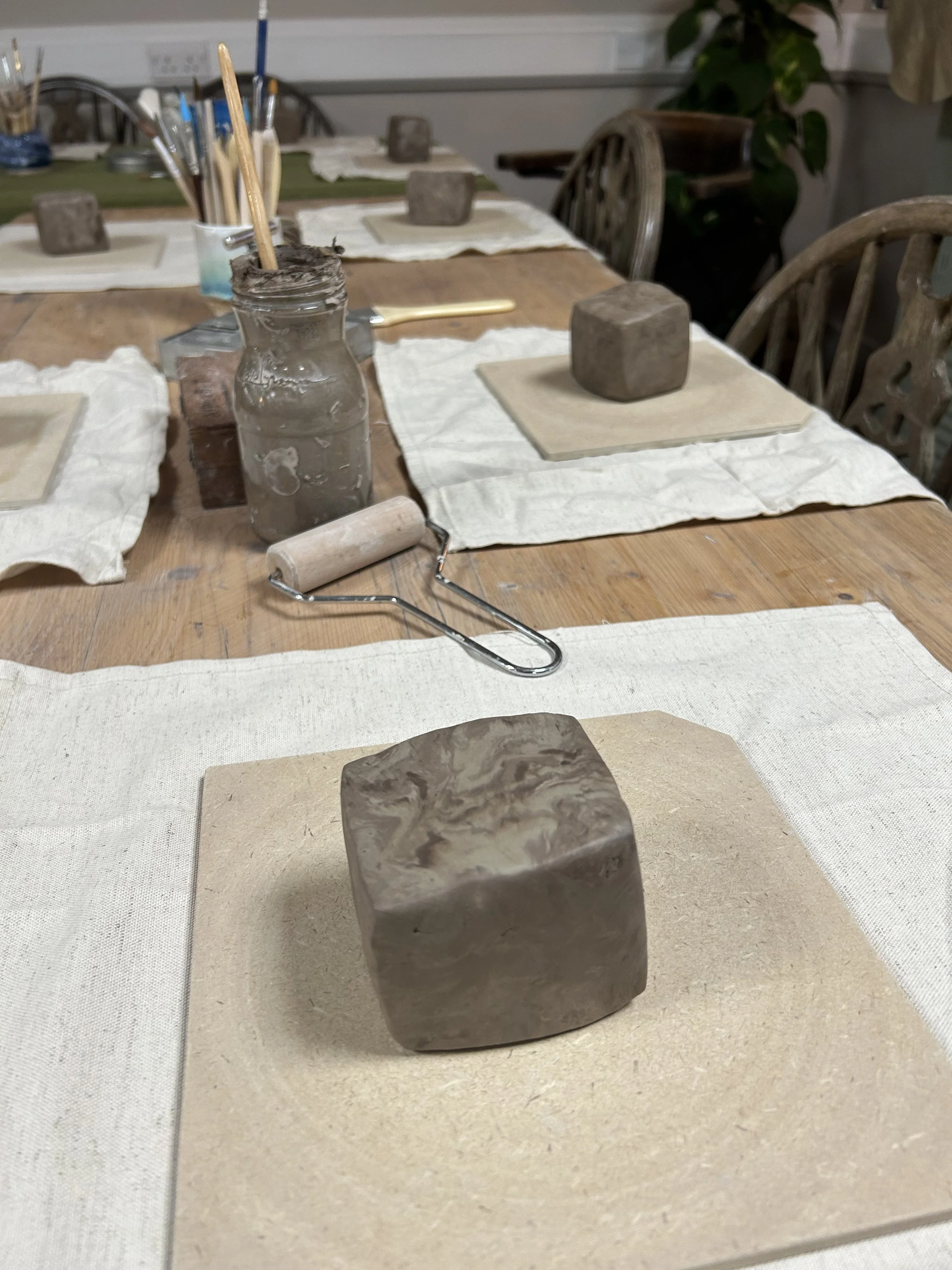 A square block of clay on a beige board at a pottery class setup, with other similar blocks and tools on a wooden table.