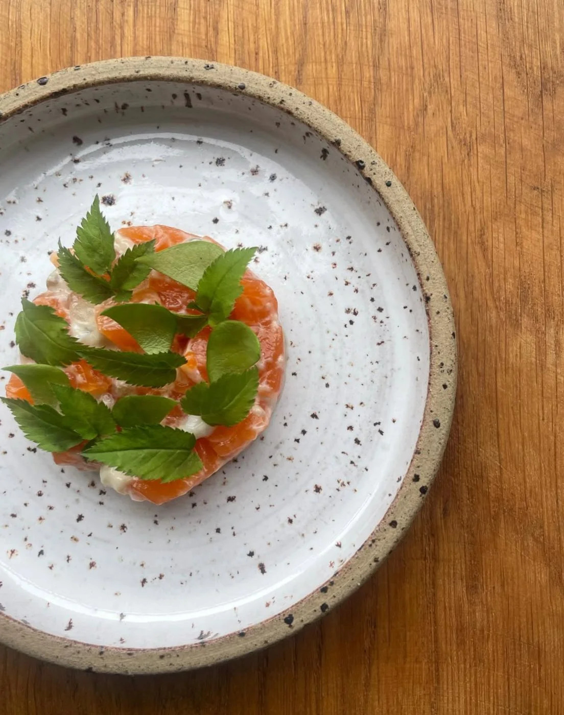 Sliced salmon tartare topped with green herbs on a white plate with a speckled rim, placed on a wooden surface.