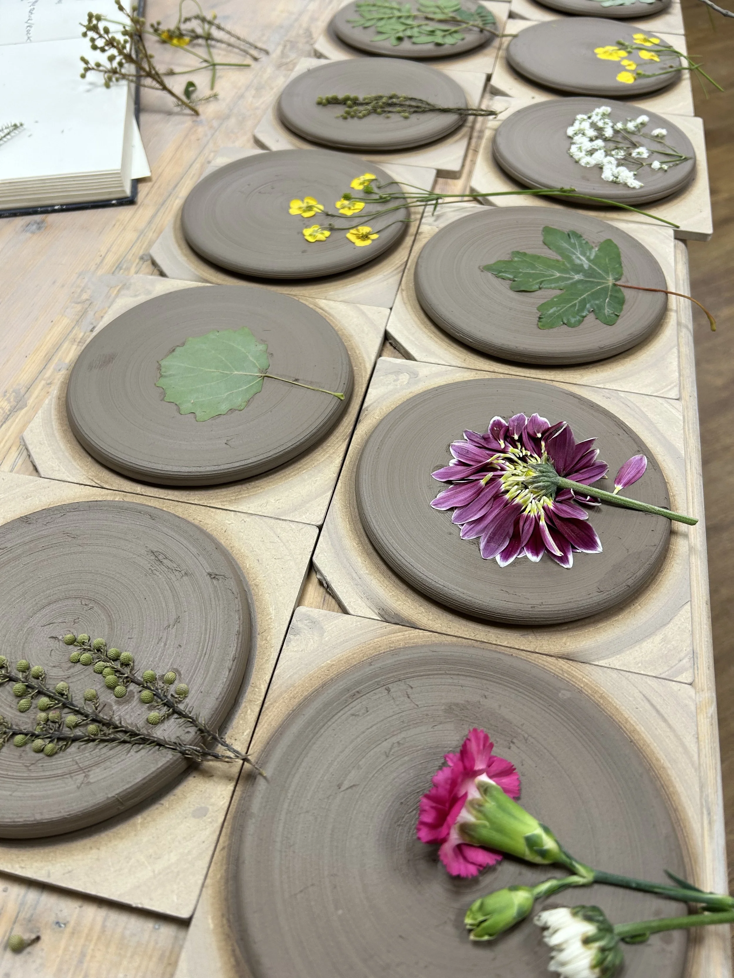 Several gray and beige ceramic plates with different colorful flowers and leaves on top, arranged on a wooden table.