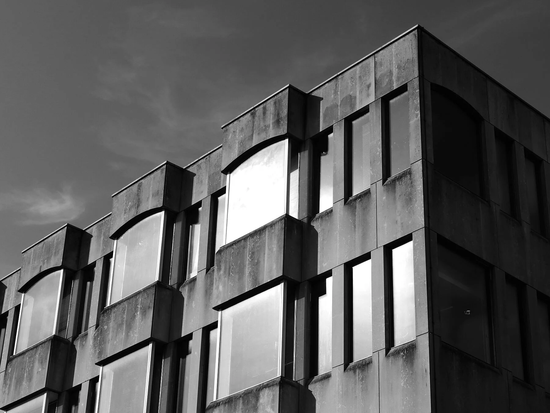 Black and white photo of a modern building with large windows and textured concrete walls, under a cloudy sky.