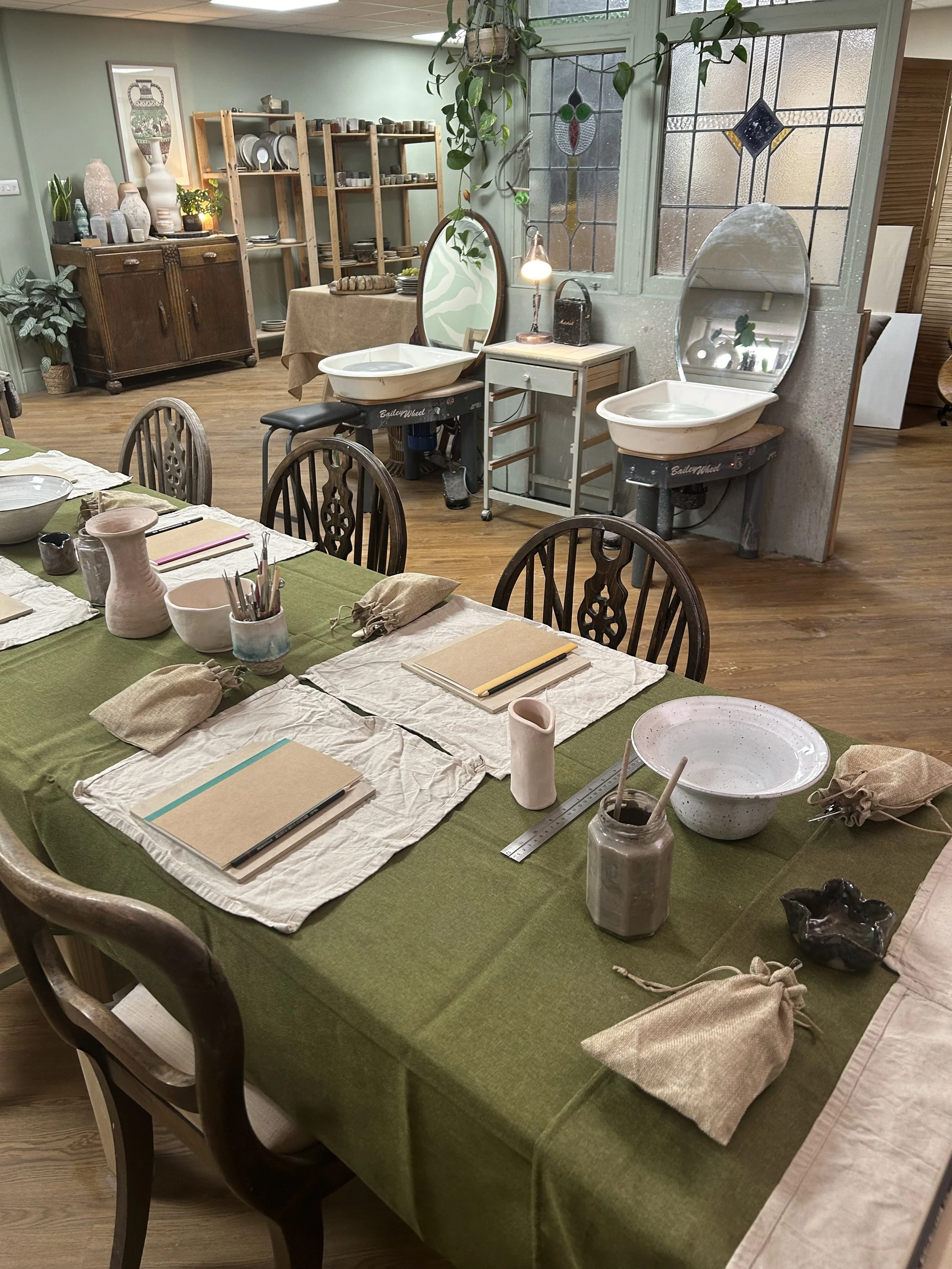 A room set up for a pottery or art class, featuring a long table with green and white tablecloths, art supplies, notebooks, and napkins. In the background are pottery wheels, mirrors, shelves with pottery, and decorative vases, with stained glass win