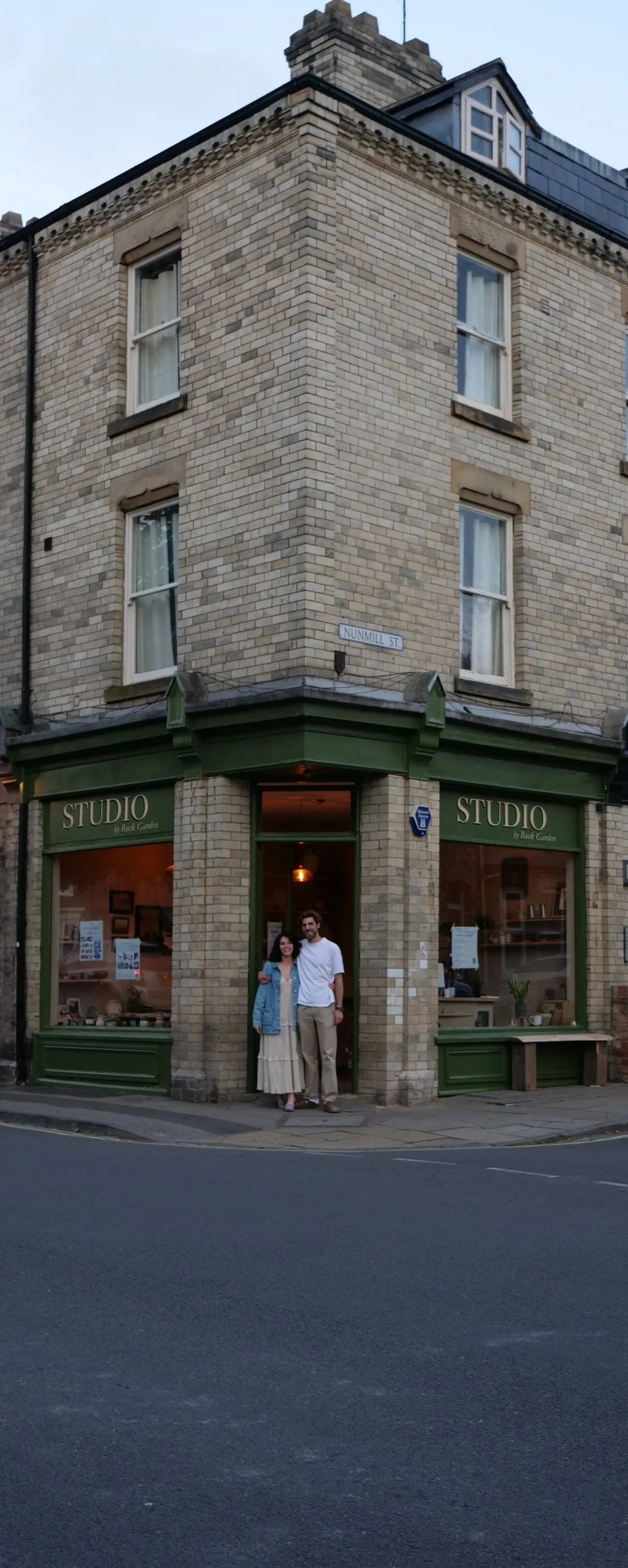 A couple standing outside a corner building with a green storefront called 'STUDIO by Raquel Gorria,' on Nummil Street, with a three-story brick structure. The woman is wearing a beige skirt and jacket, and the man is wearing a white shirt and beige pants.
