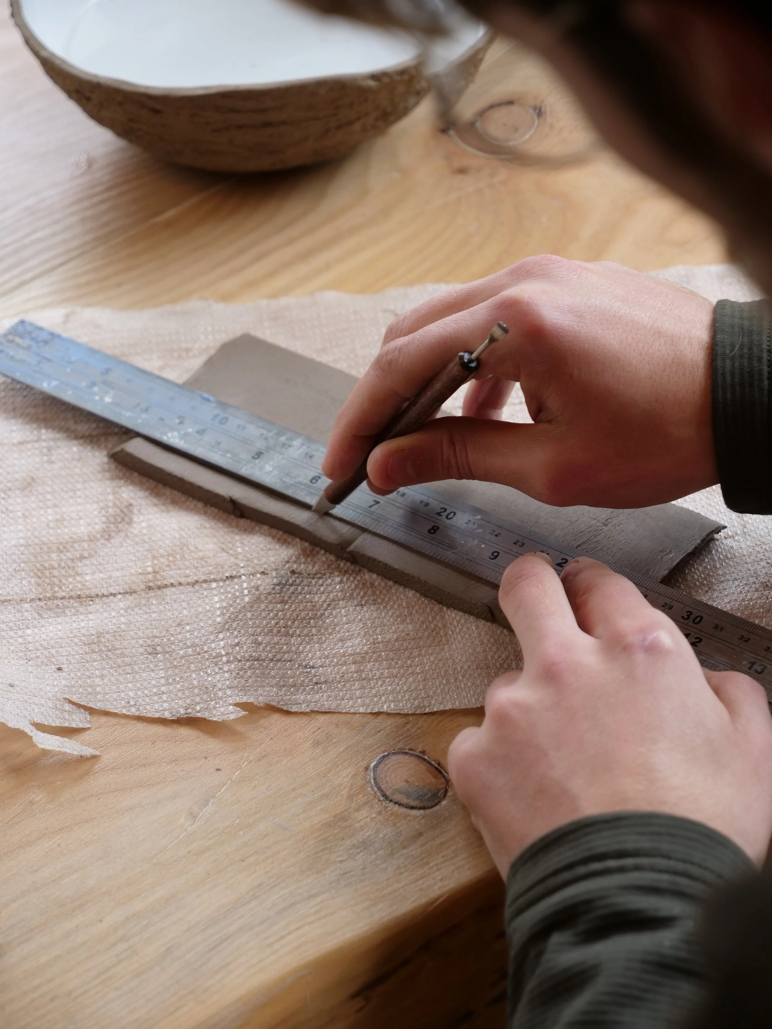 Person using a metal tool to carve or cut a piece of clay or similar material on a wooden table, with a ruler and a bowl in the background.