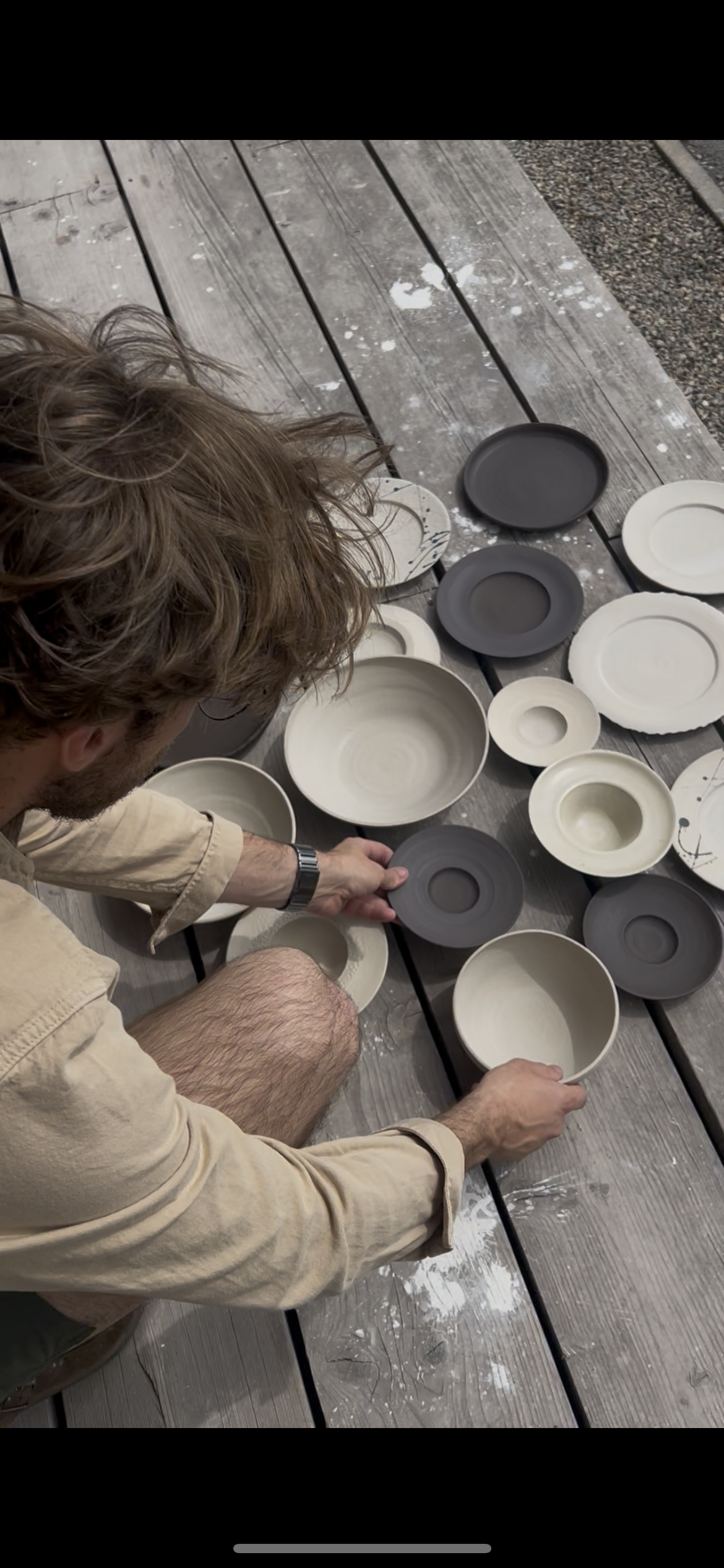 A person arranging ceramic bowls and plates on a wooden surface outdoors, with some pottery being black and others cream-colored.