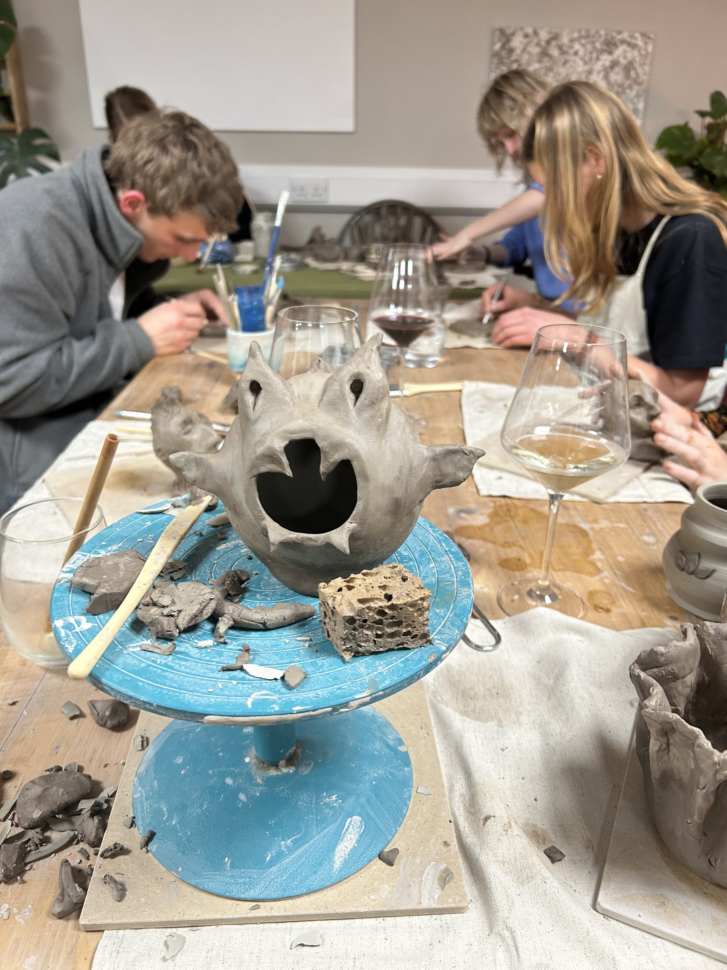 People working on ceramic sculptures at a pottery class, with clay and tools on a wooden table, and a sculpture of a creature with a surprised expression in the foreground.