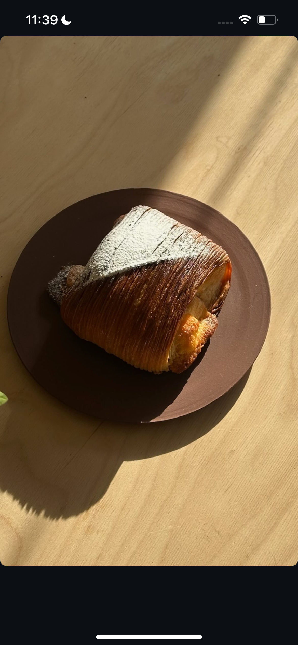 A slice of apple pie dusted with powdered sugar on a dark brown plate, placed on a light wooden surface.