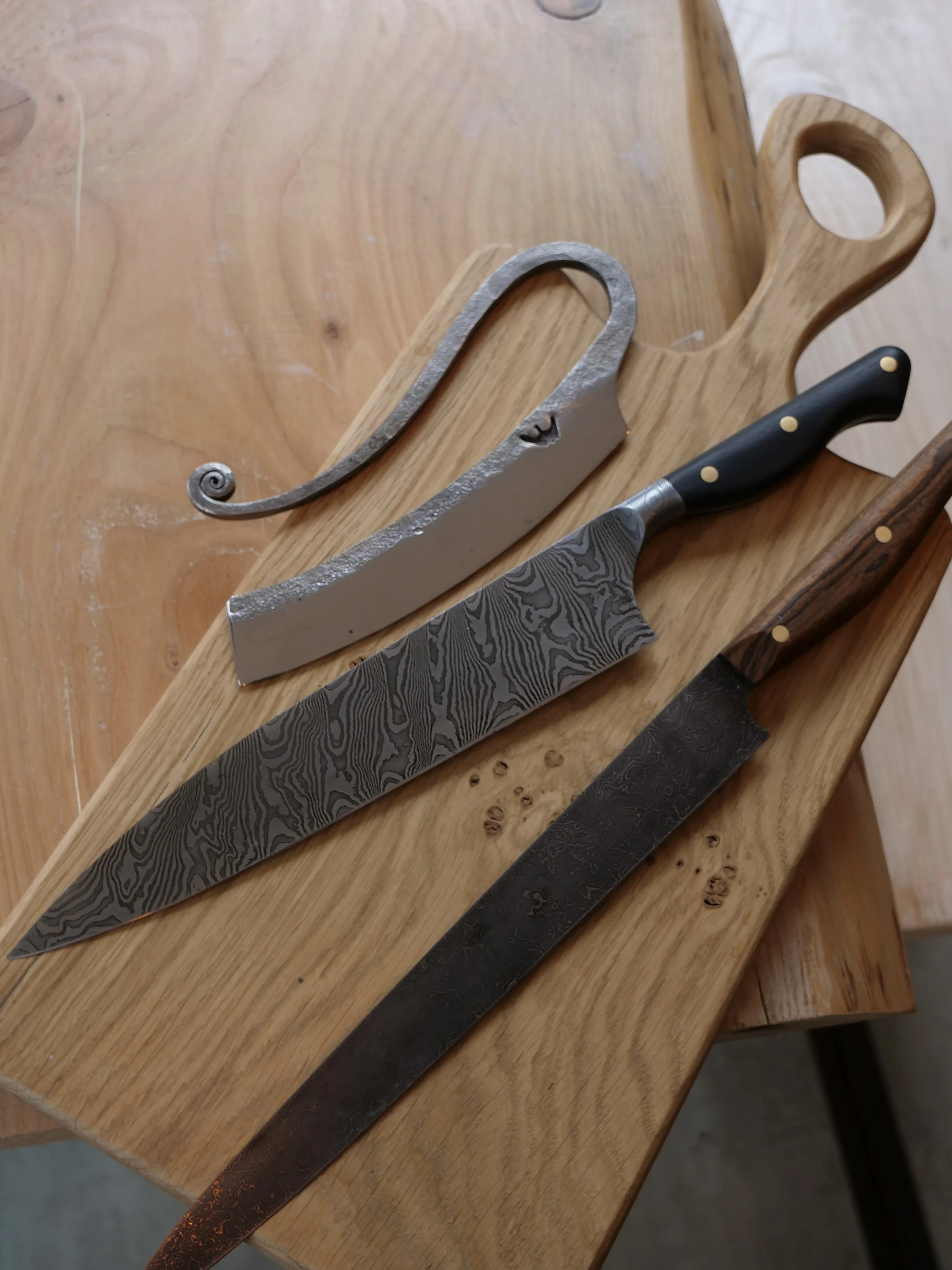 Three kitchen knives of different styles on a wooden cutting board with a handle. The first knife has a Damascus steel blade with a black handle, the second is a large curved chef's knife with a grey handle, and the third is a long, narrow steel blade with a wooden handle.