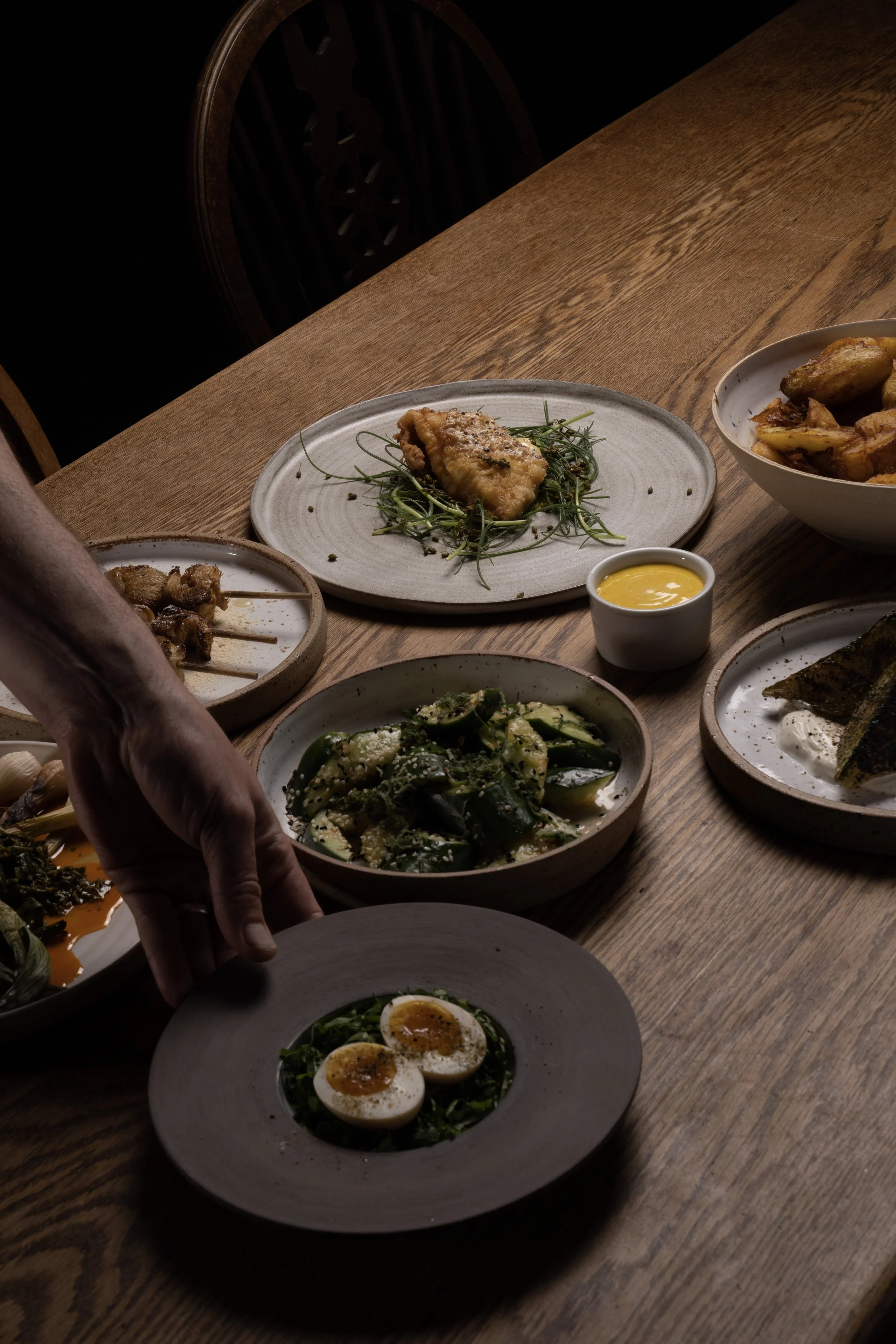 Assorted dishes on a wooden table, including a plate of halved soft-boiled eggs garnished with herbs, cucumber salad, fried chicken, potato wedges, grilled fish with sauce, and skewers.