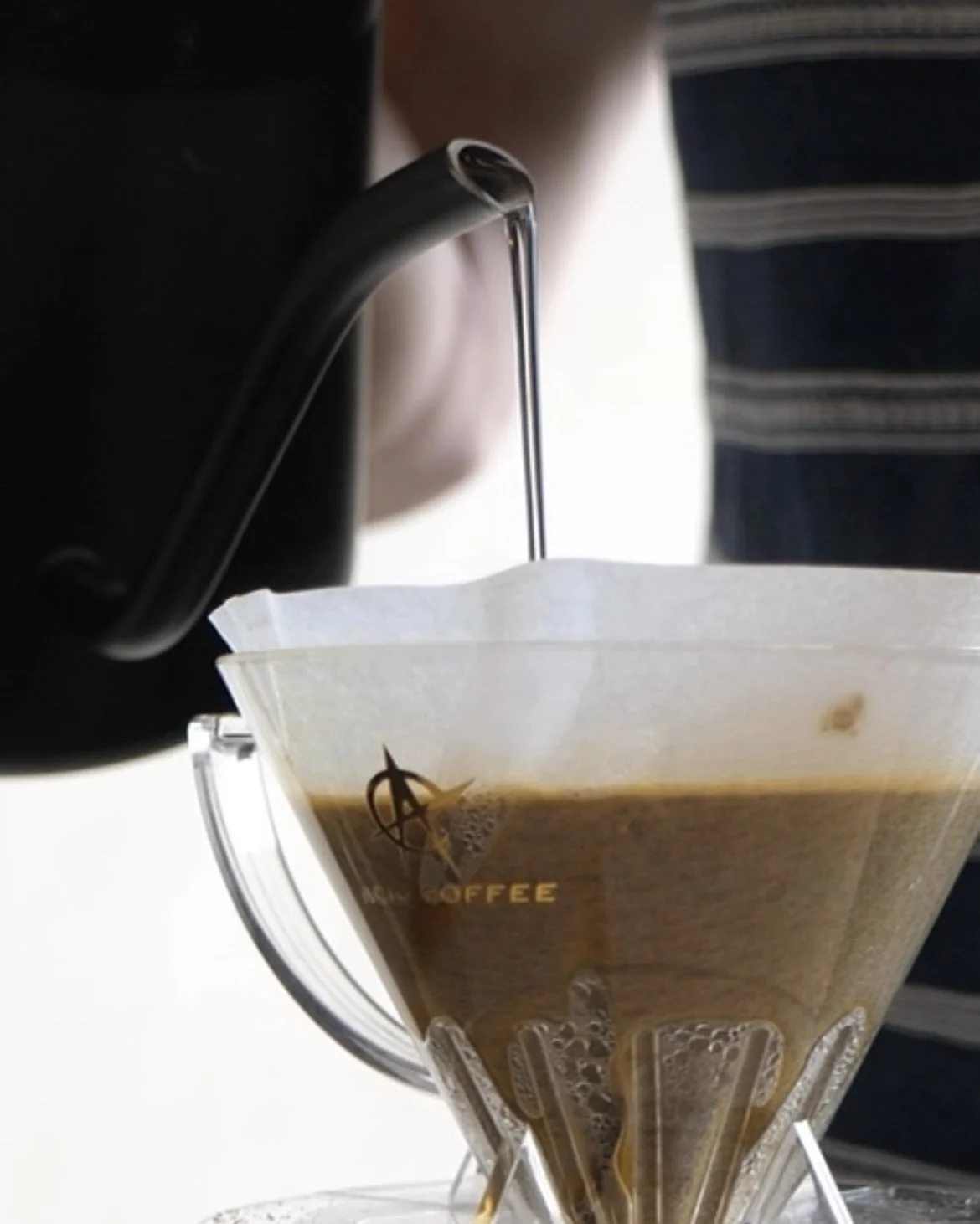 Close-up of hot coffee being brewed through a pour-over coffee maker with a metal gooseneck kettle pouring hot water into a clear plastic cone-shaped filter filled with coffee grounds.