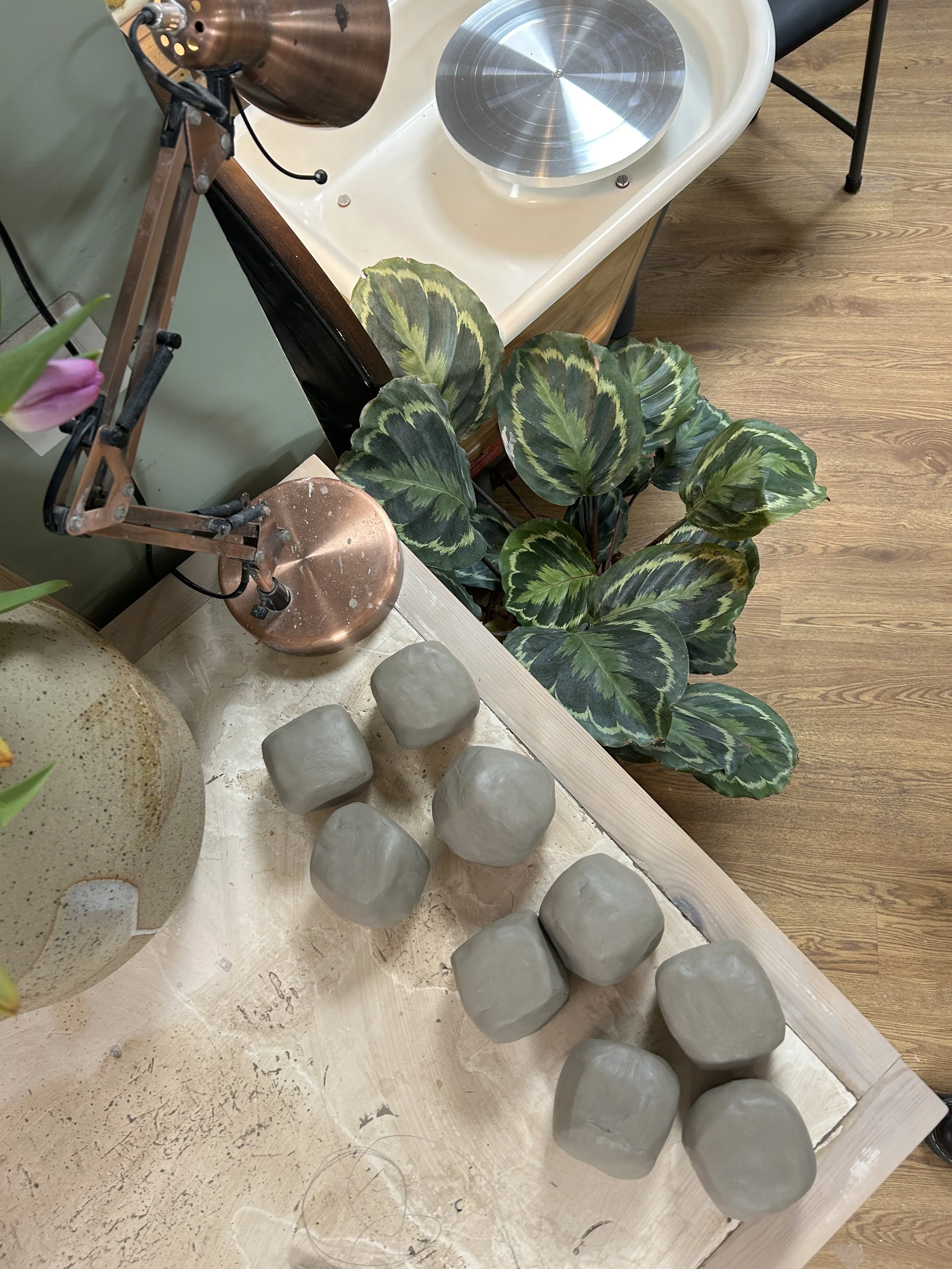 A wooden table with eight small gray clay sculptures, a large decorative vase, a green leafy plant, a copper desk lamp, and a small ceramic sculpture of a yellow flower.