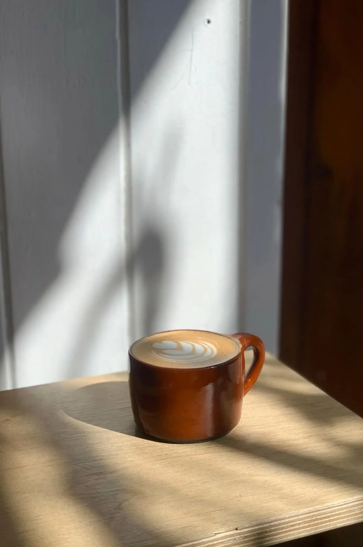 A brown ceramic cup filled with latte art coffee on a wooden table with sunlight and shadow patterns.