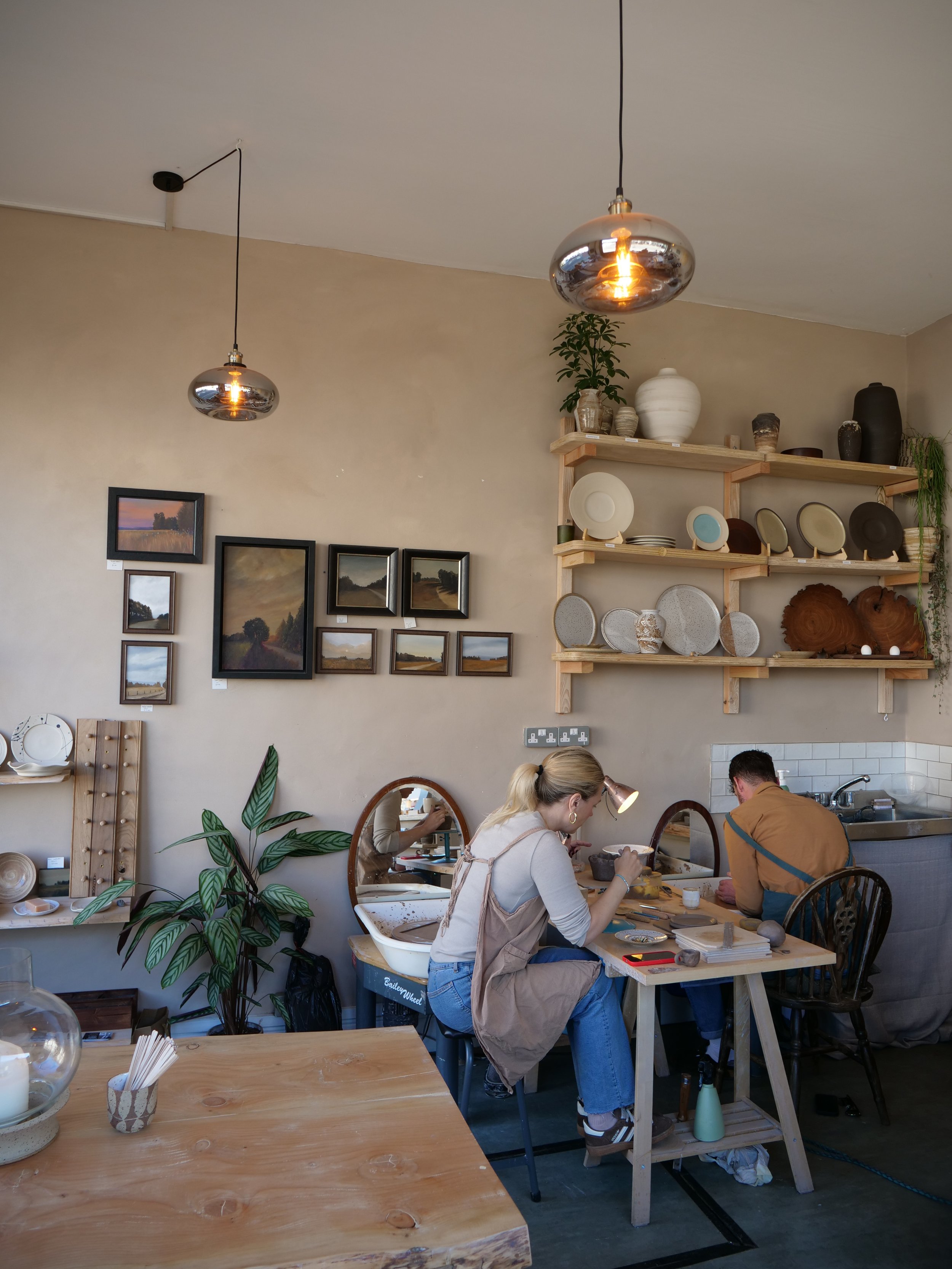 Two people working on clay pottery at a table in a cozy, art-studio-style space with shelves of pottery and wall art.