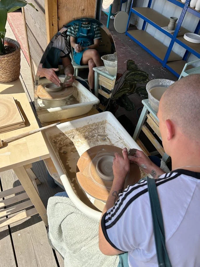 A person with a shaved head is pottery making on a wheel, with a mirror in front reflecting their activity and another person sitting behind them. The workspace has pottery tools, bowls, and shelves with ceramic pieces.