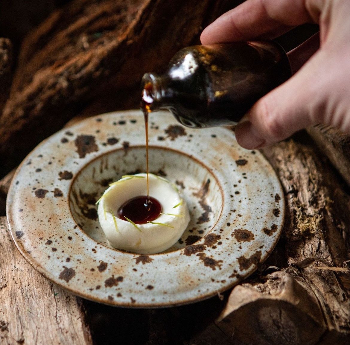 A hand drizzling dark syrup over a round white cheese or dessert on a rustic, speckled ceramic plate, with a wood log background.