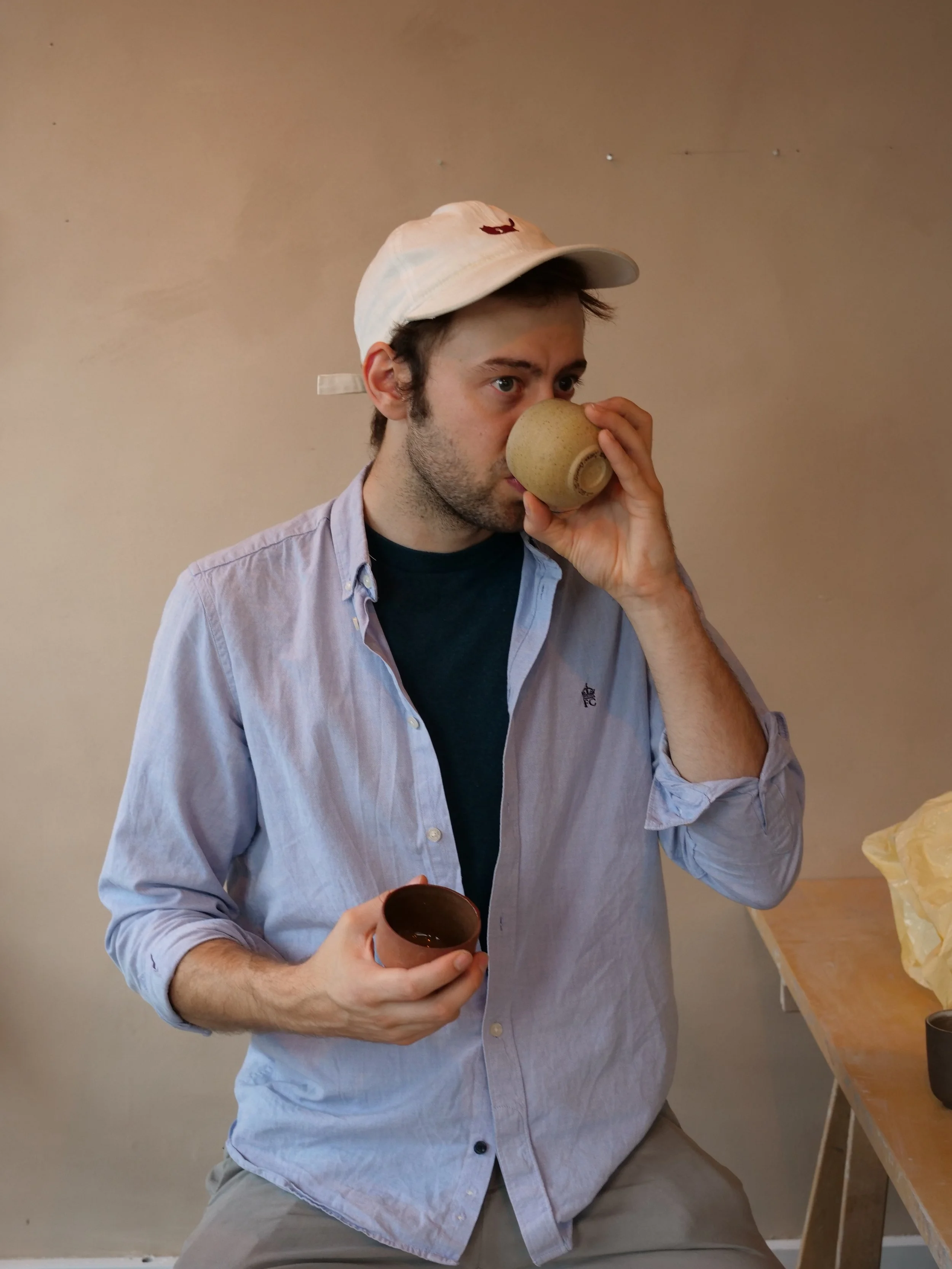 A young man sipping from a ceramic cup while holding another in his left hand, wearing a white cap, light blue shirt, and dark T-shirt underneath, standing near a wooden table with paper and cups.
