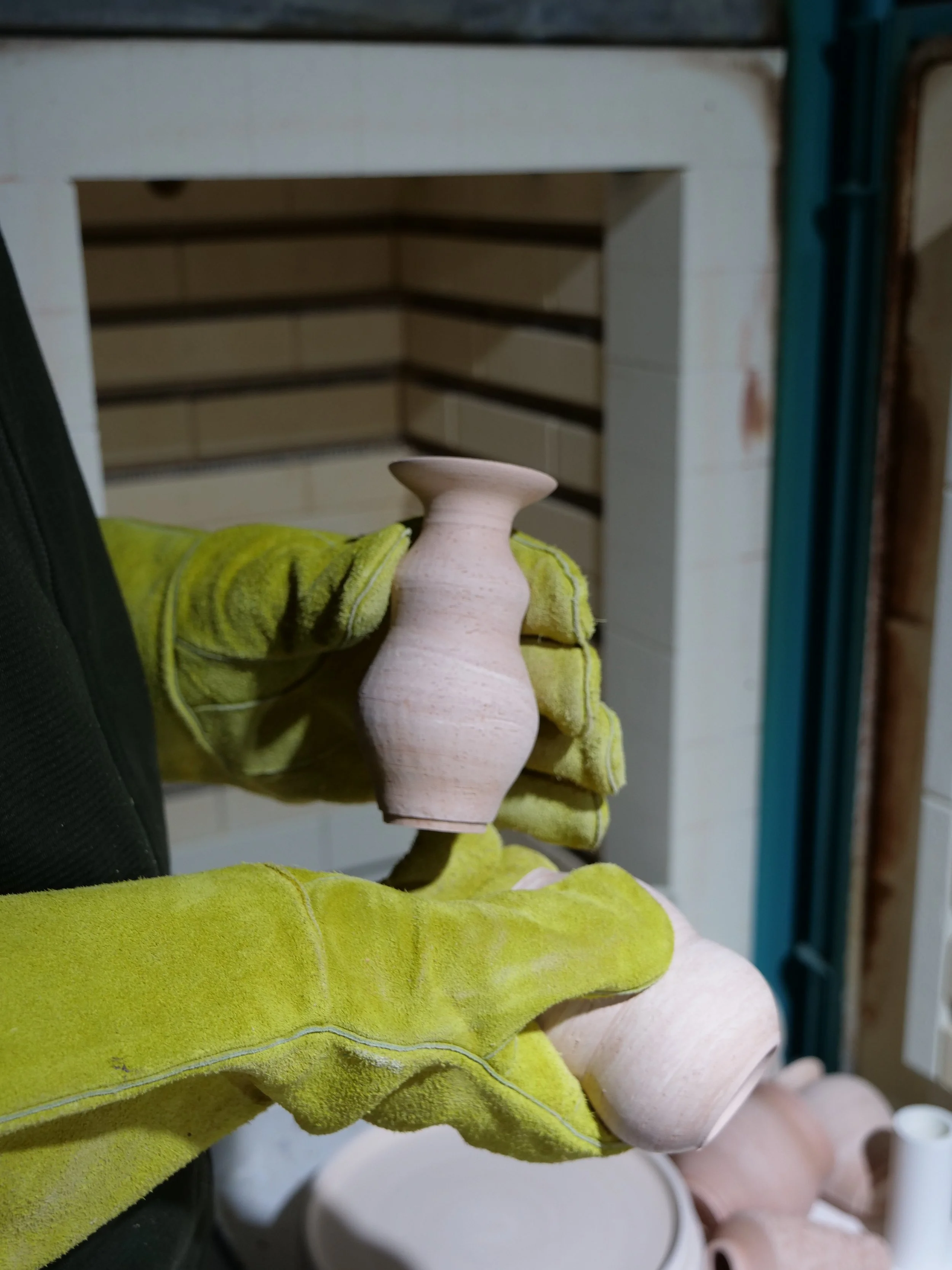 A person wearing yellow gloves holding unfinished pottery, with pottery items on a table in the background.