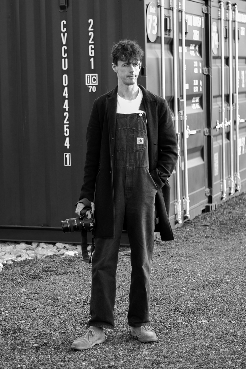 A young man standing outdoors on gravel, holding a camera with a strap, in front of shipping containers, wearing a long coat, overalls, a t-shirt, and boots, in black and white.