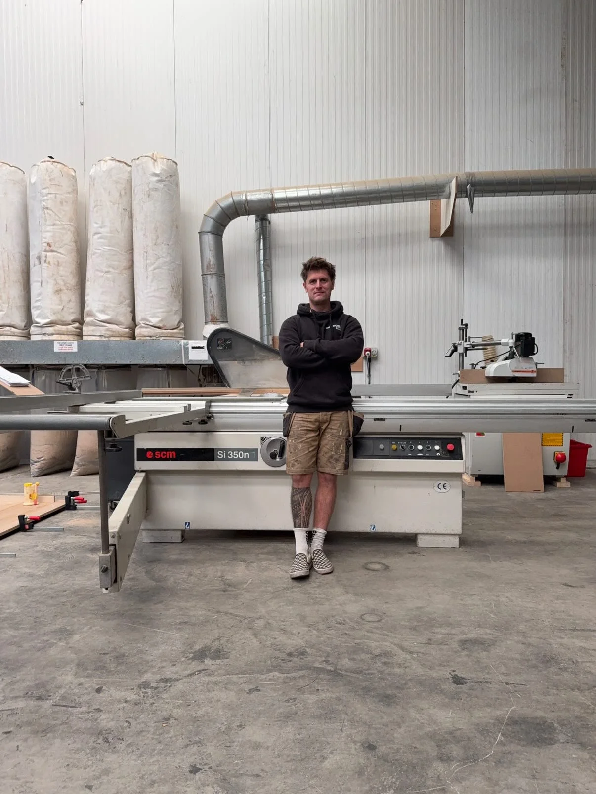 Man standing with arms crossed in a woodworking or manufacturing workshop, surrounded by large machinery and equipment.