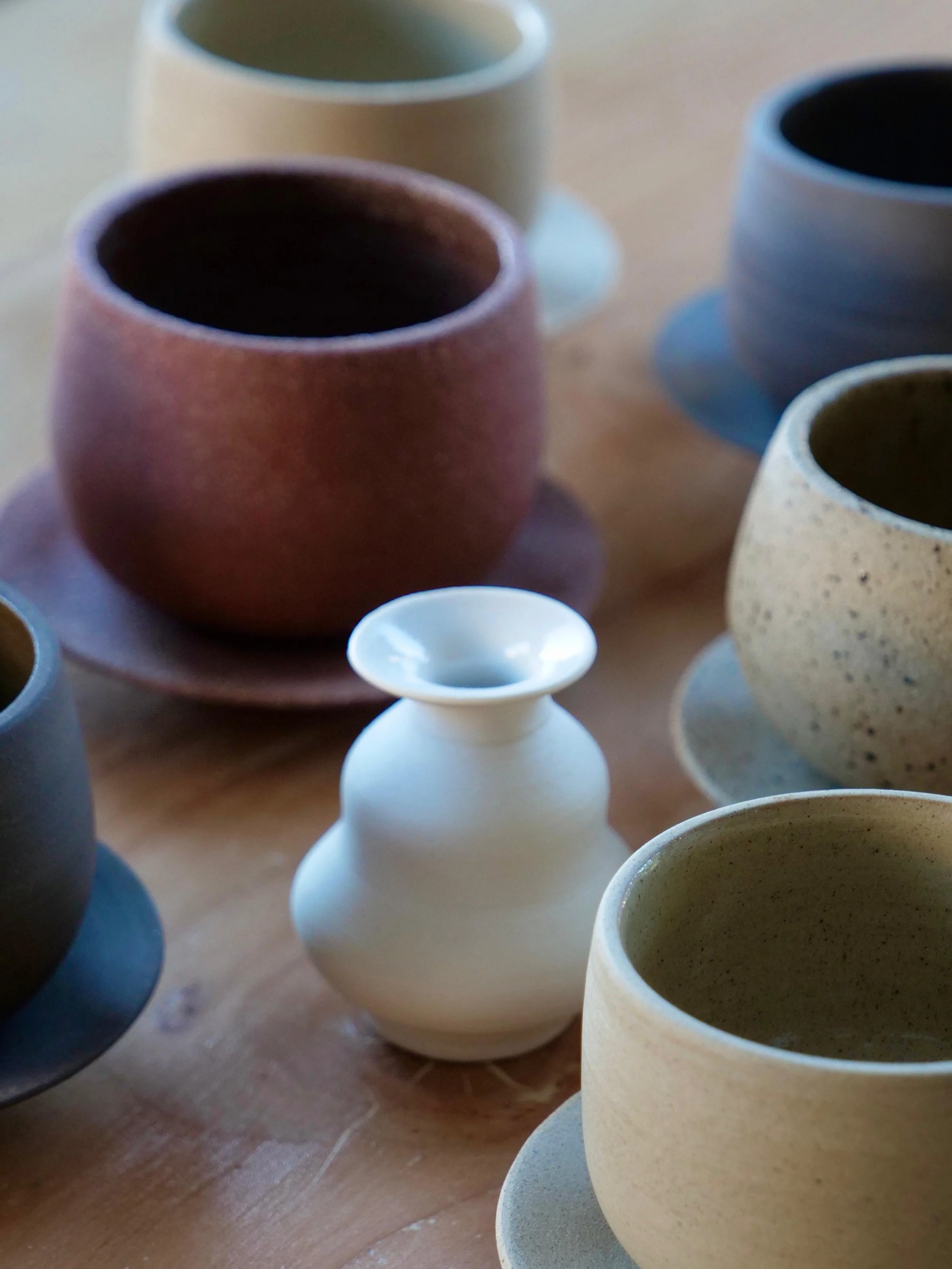 A collection of ceramic cups and a small white vase on a wooden table.