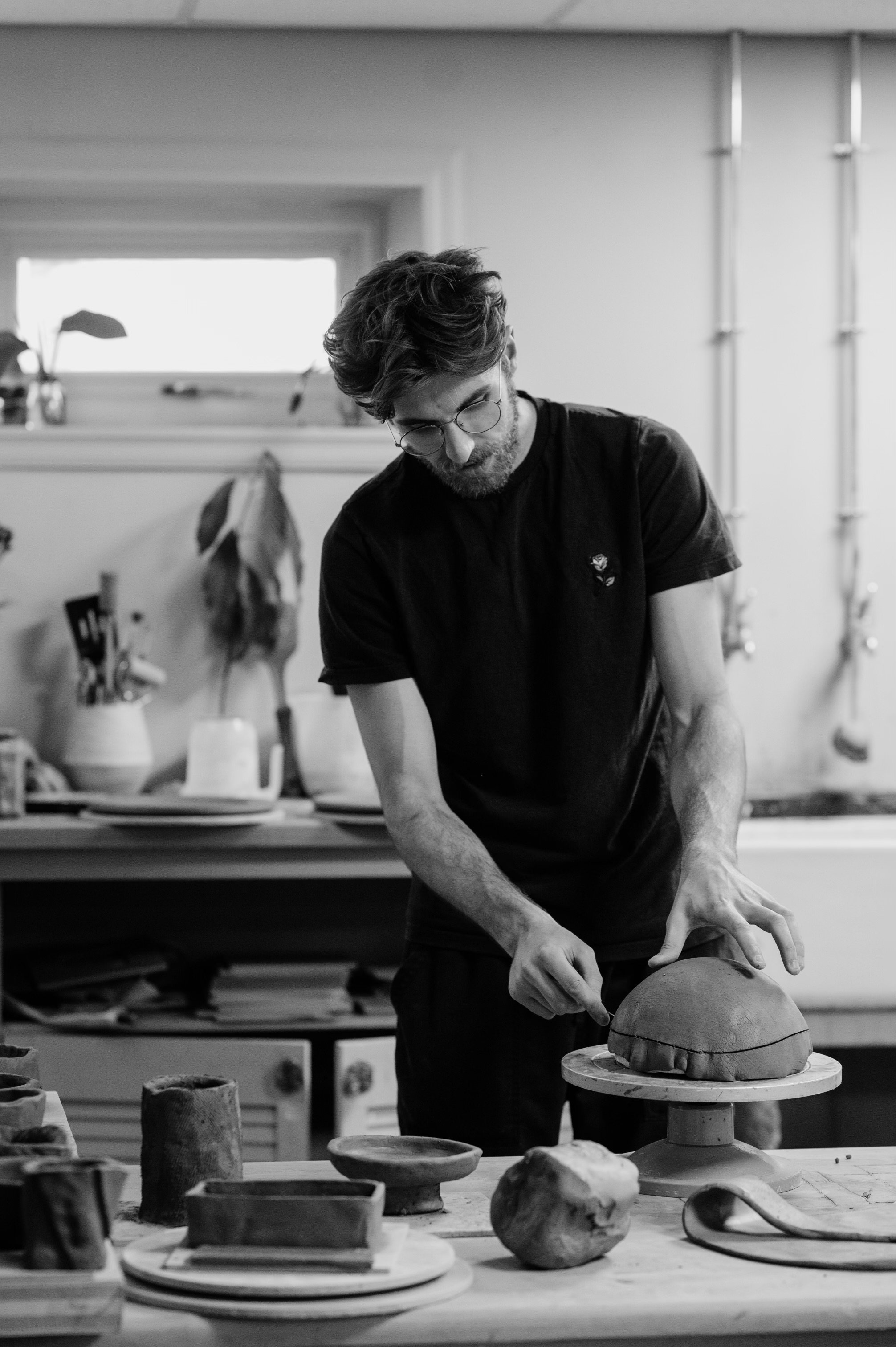 A man with glasses and a beard is working on a ceramic piece in a studio with pottery tools and unfinished pottery on the table.