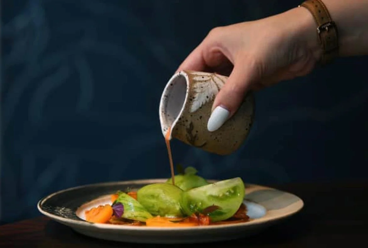 A hand with white nail polish and a brown wristwatch pouring sauce from a small cup onto a plate of salad with green vegetables and colorful garnishes