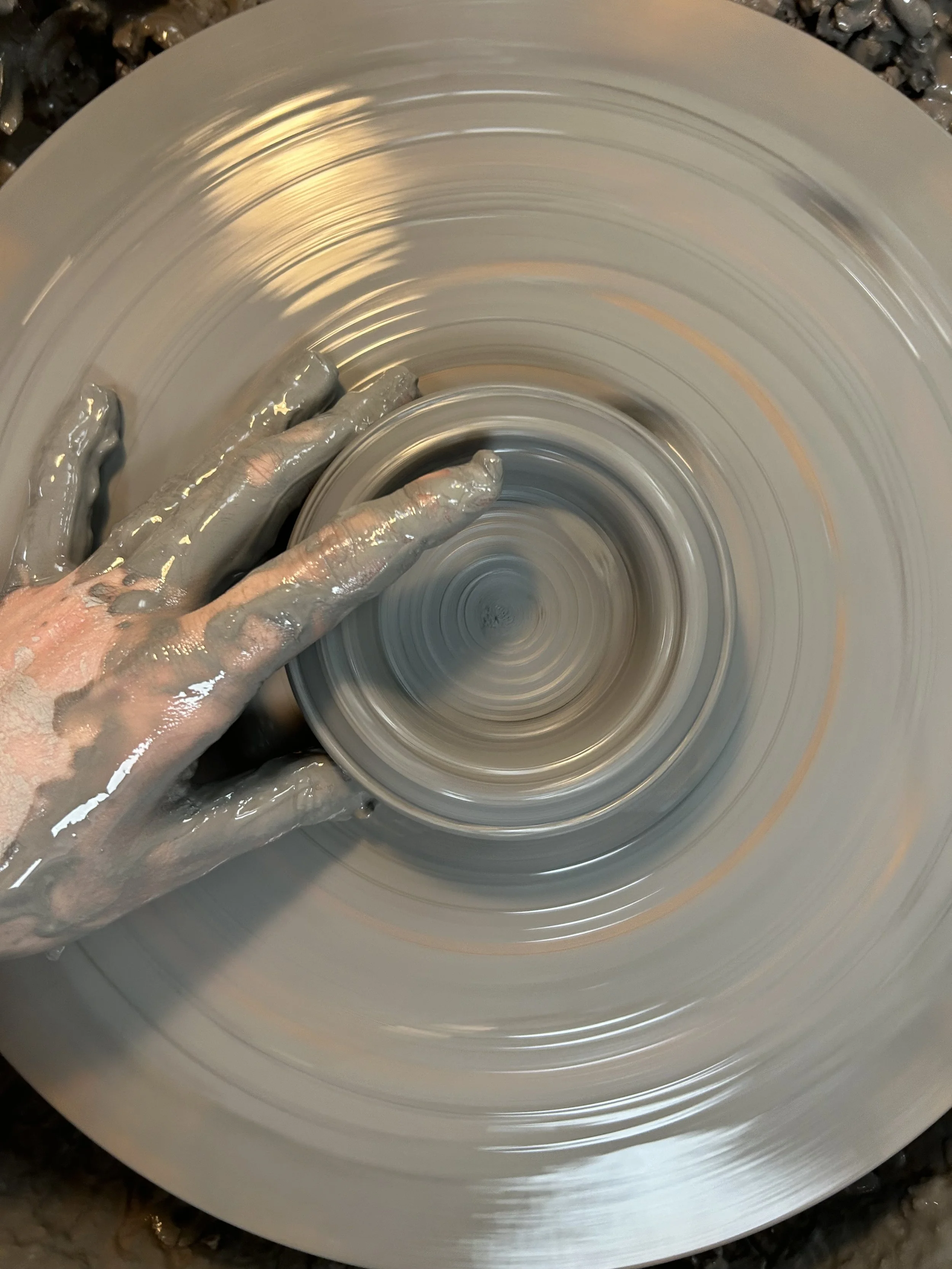 A person’s hand covered in wet clay shaping a pot on a spinning pottery wheel.