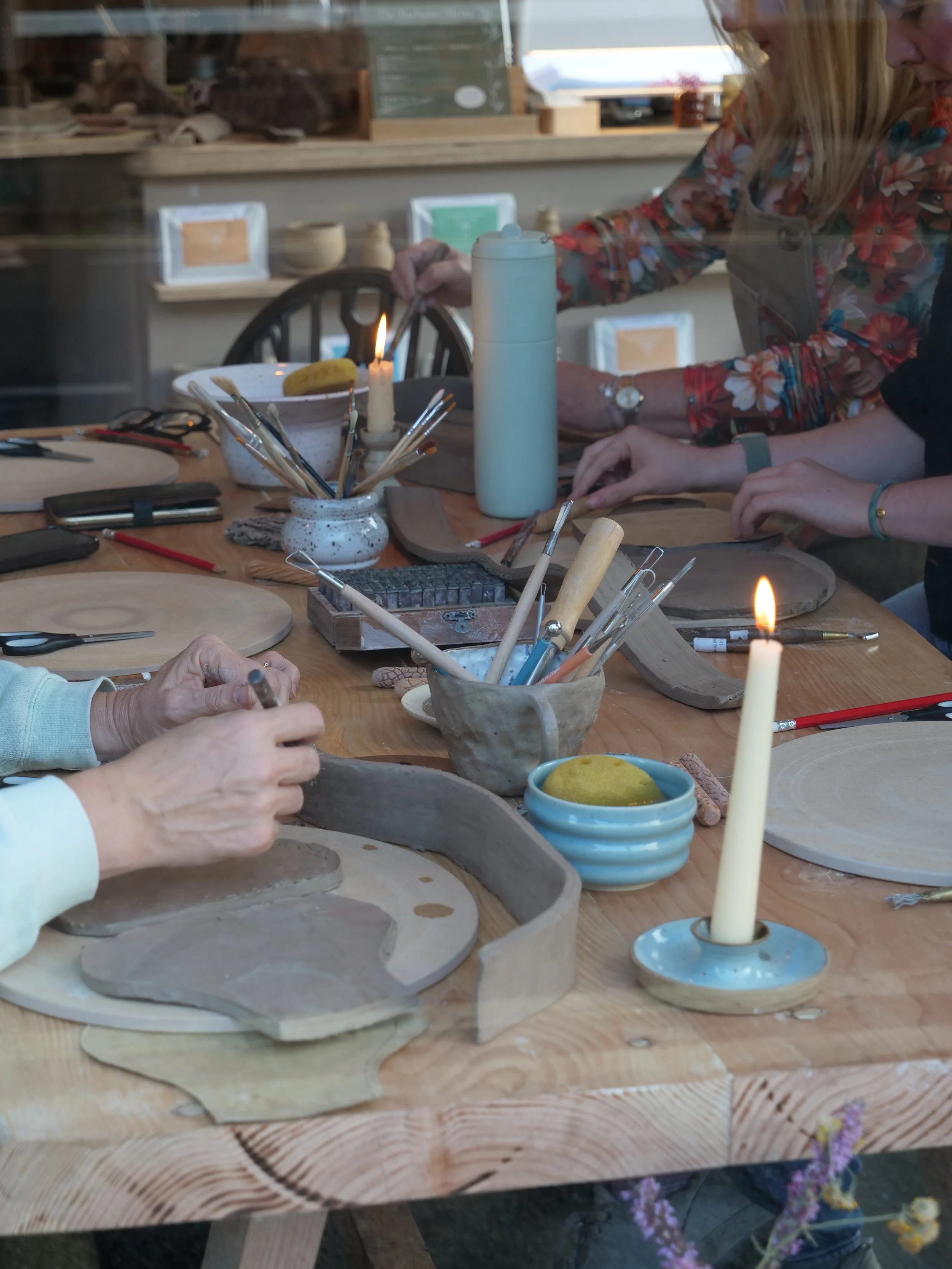 People working on a pottery project at a wooden table with clay, pottery tools, and candles.