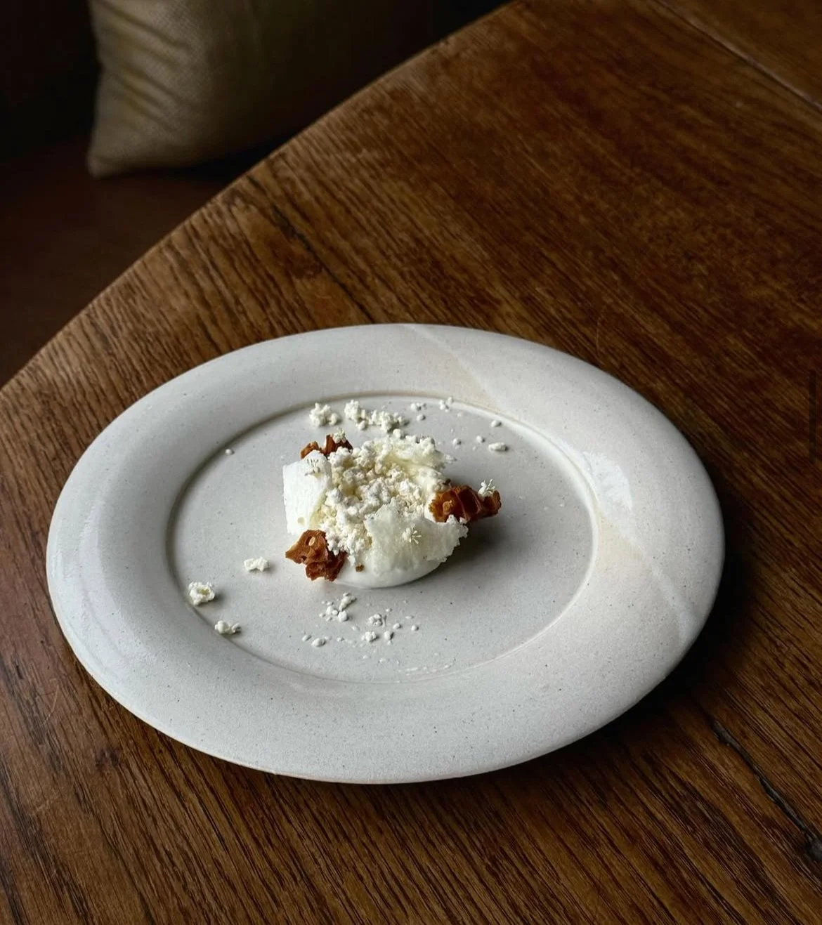 A dessert with white cream, crumbled white cheese, and small pieces of caramelized nuts on a white oval plate set on a wooden table.