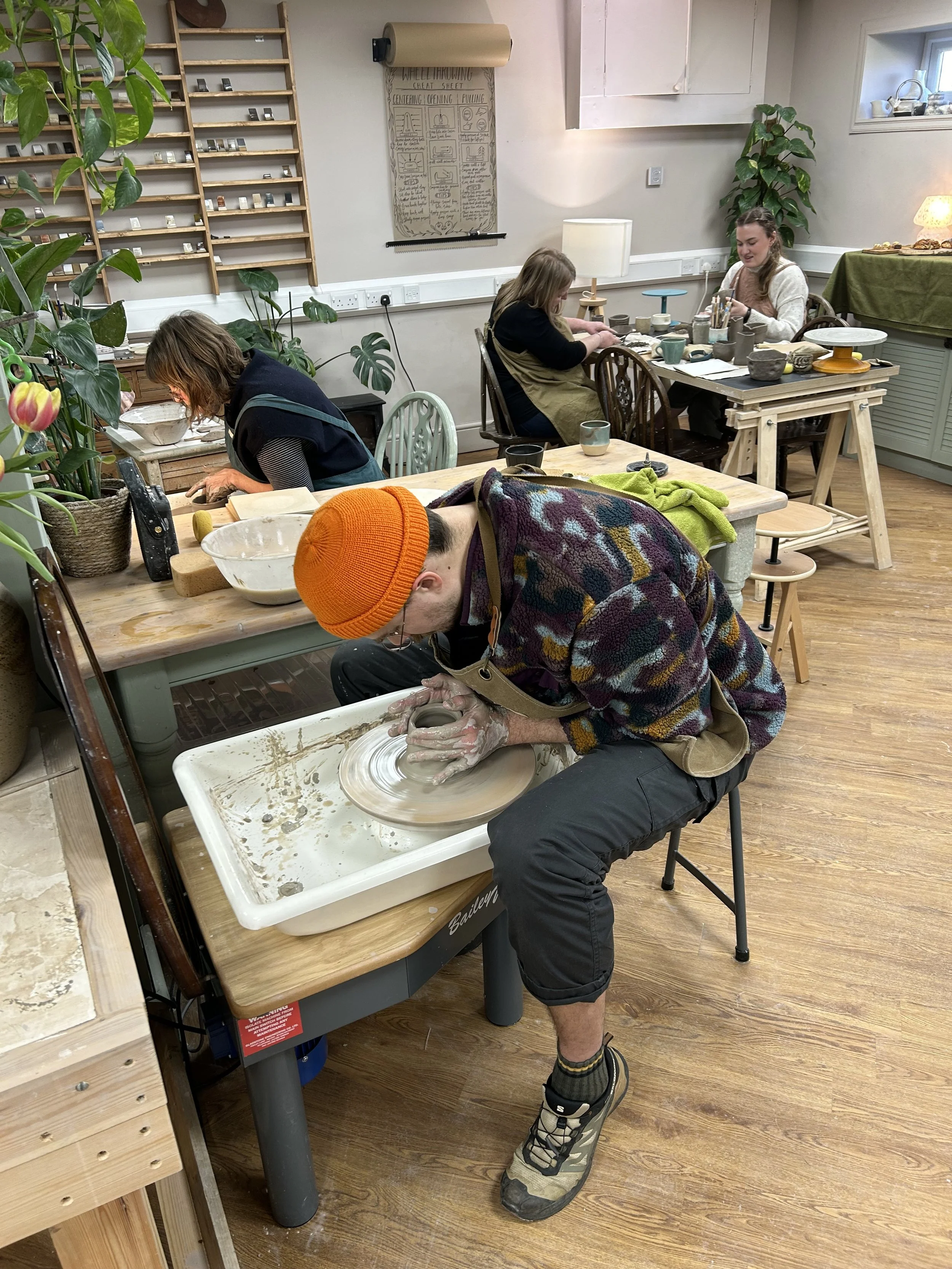 A man wearing an orange beanie and colorful sweater is shaping pottery on a wheel in a pottery studio. Three women are working at tables in the background, engaged in pottery or arts and crafts activities.